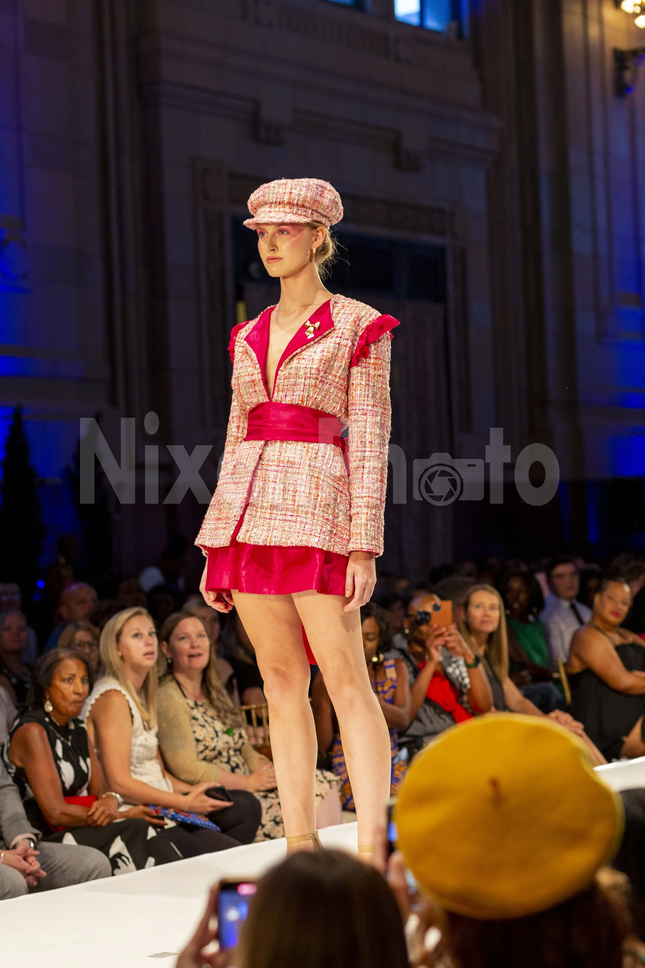 A model on a fashion runway wearing a pink and red tweed outfit with a matching hat, surrounded by an audience.