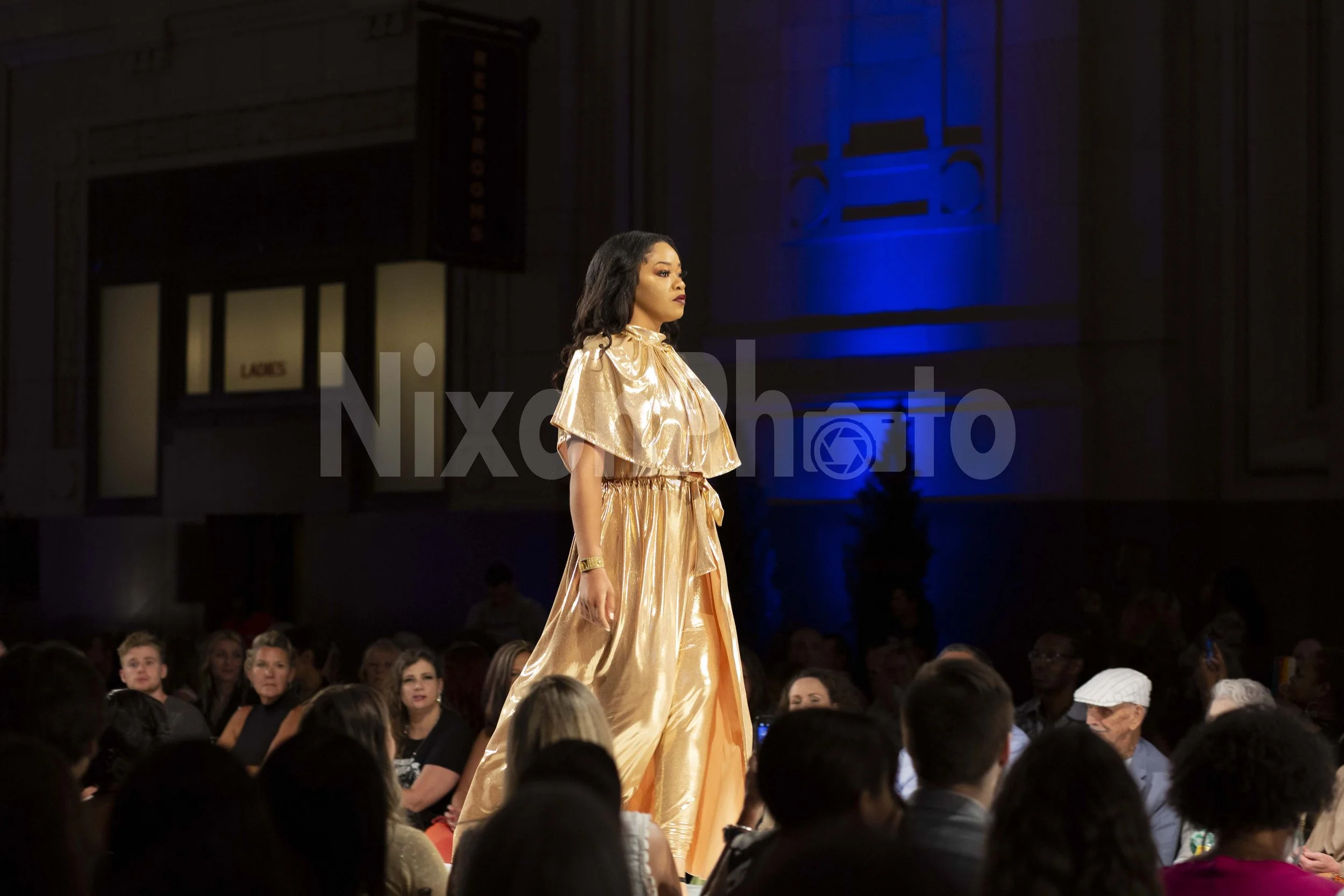 Model walking down a fashion runway in a gold dress, with an audience watching.