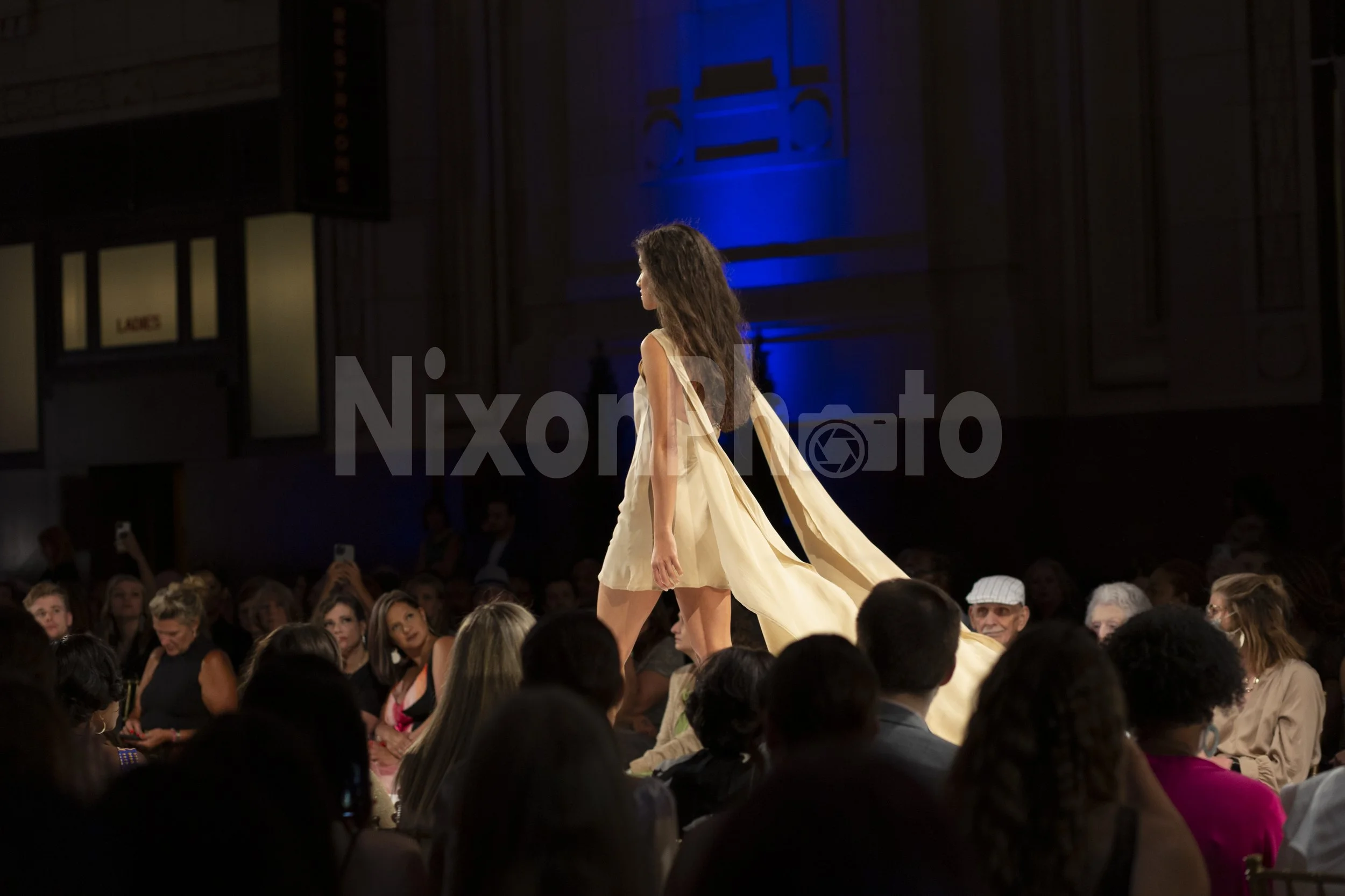 Model walking on a runway, wearing a flowing beige dress with a crowd watching at a fashion show.