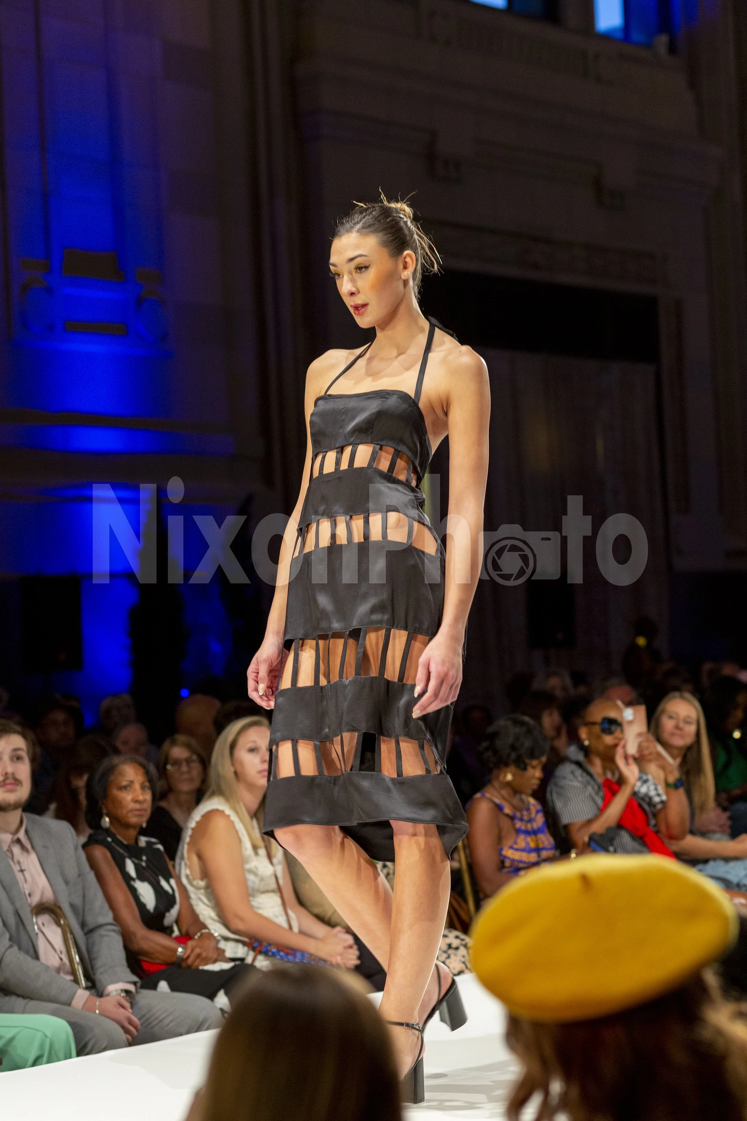 Model wearing a black cut-out dress walking on a runway during a fashion show, with an audience in the background.