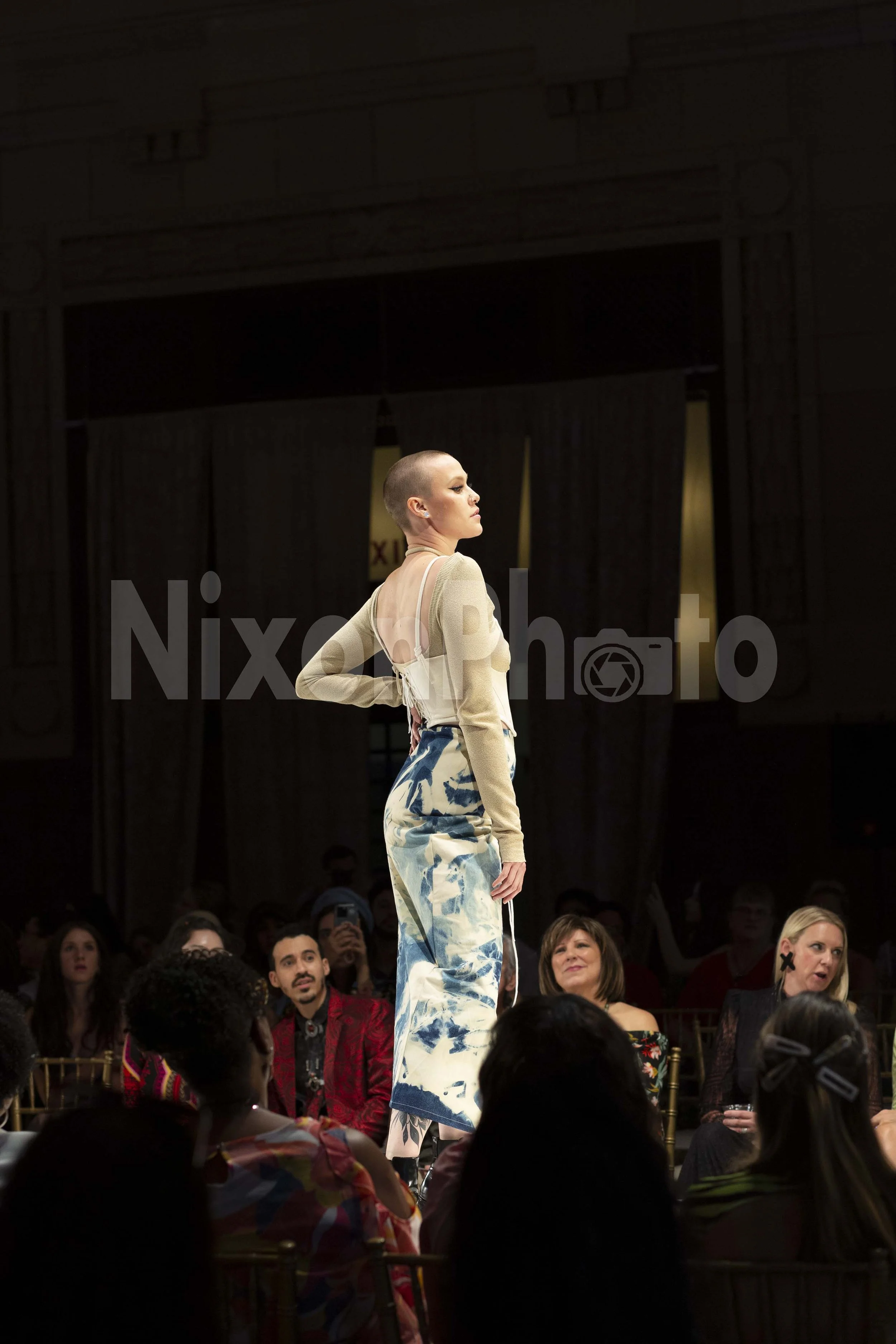 Model walking on a fashion runway wearing a long-sleeve top and blue patterned skirt, with audience watching.