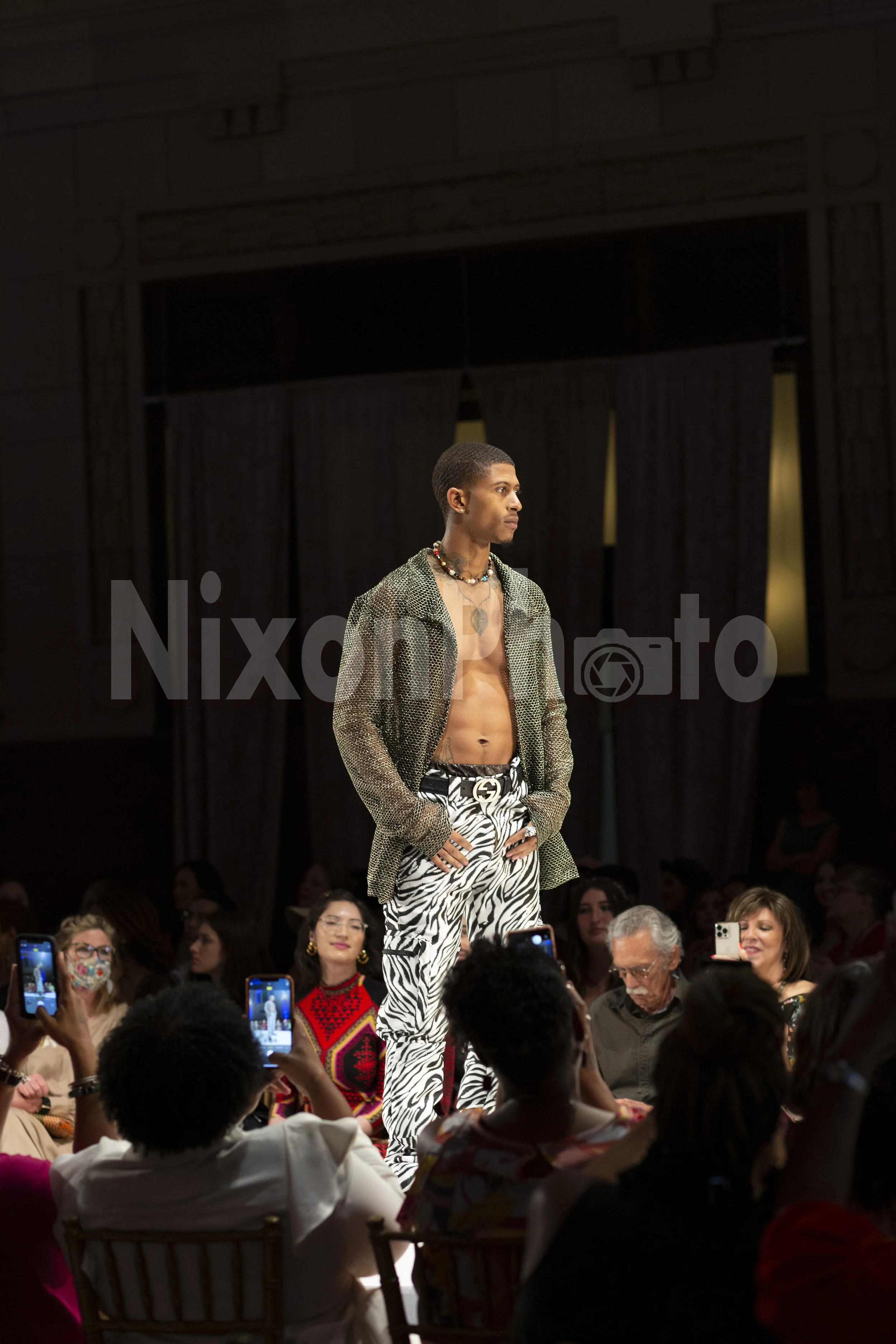 Male model walking on fashion runway wearing zebra print pants and unbuttoned shirt, surrounded by audience taking photos.