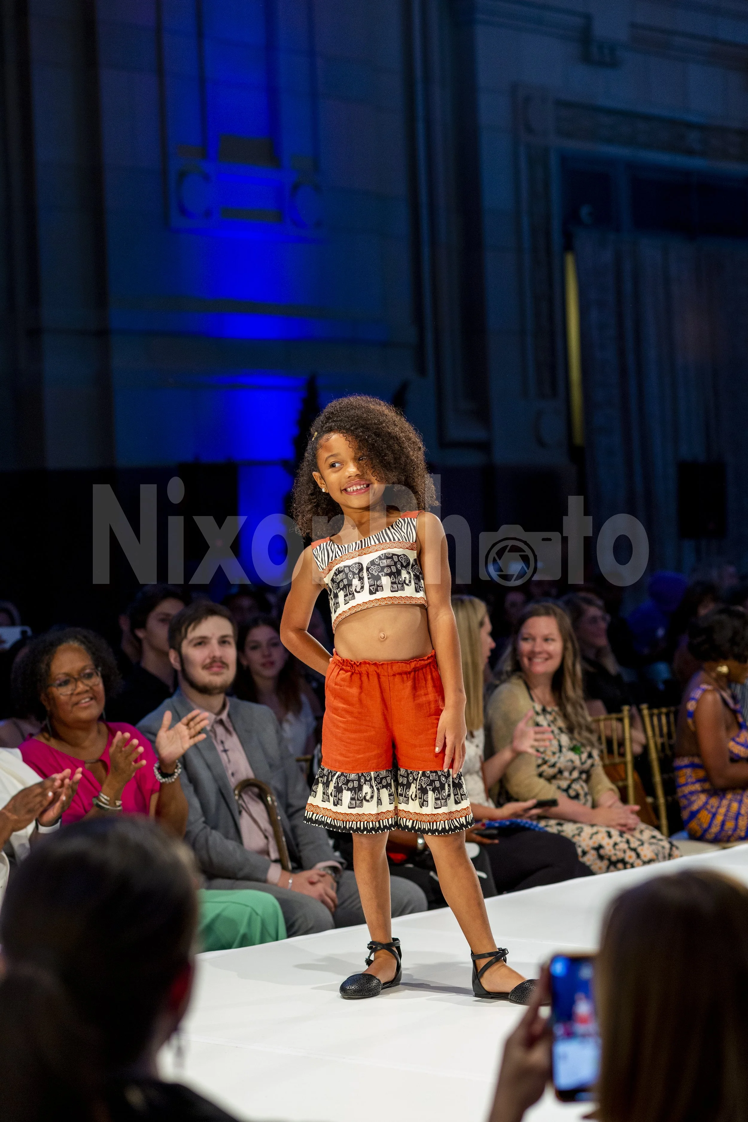 Young girl walking on a fashion show runway, wearing a matching top and shorts with an elephant print. Audience clapping in the background.