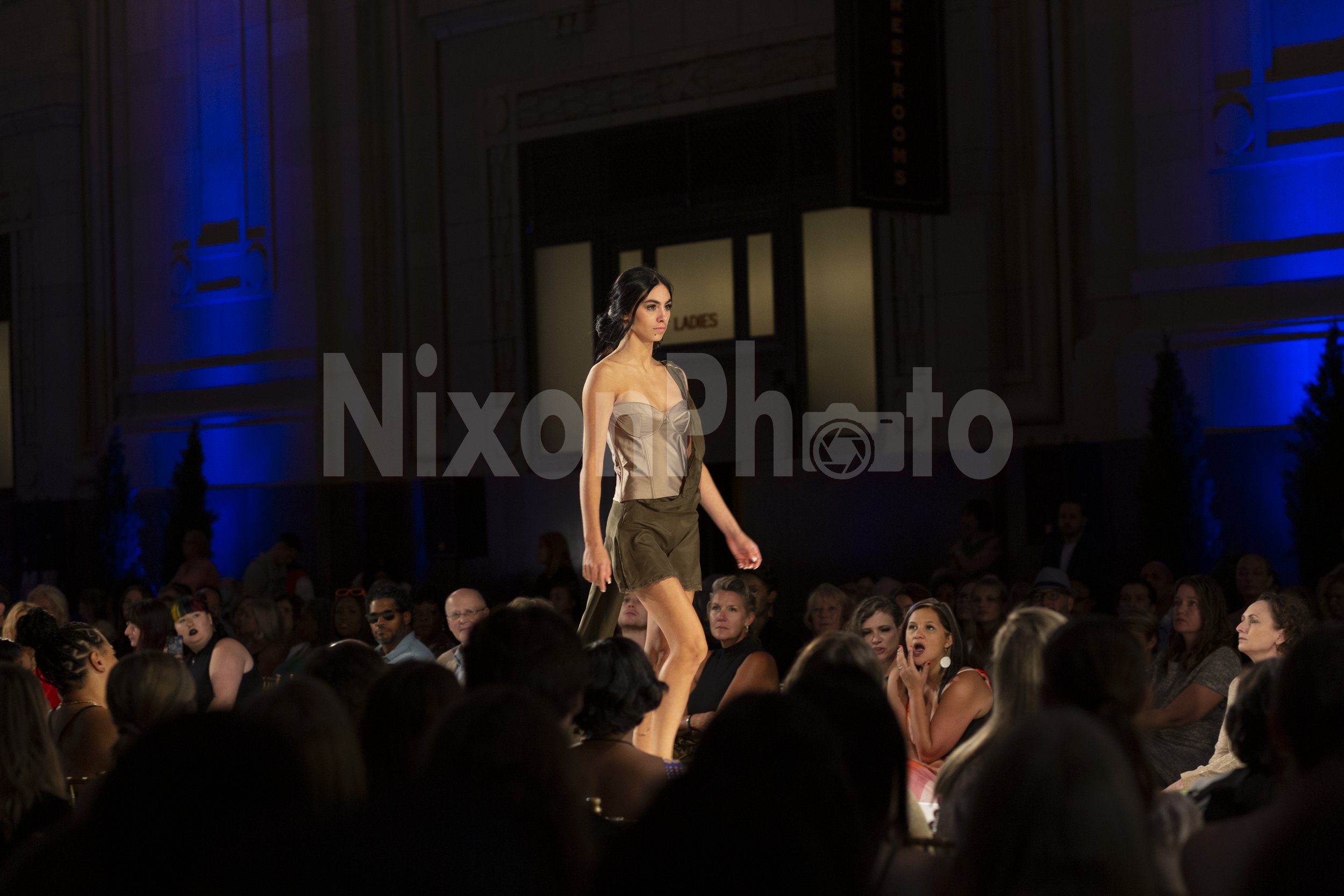 Model walking on runway at fashion show with audience watching.