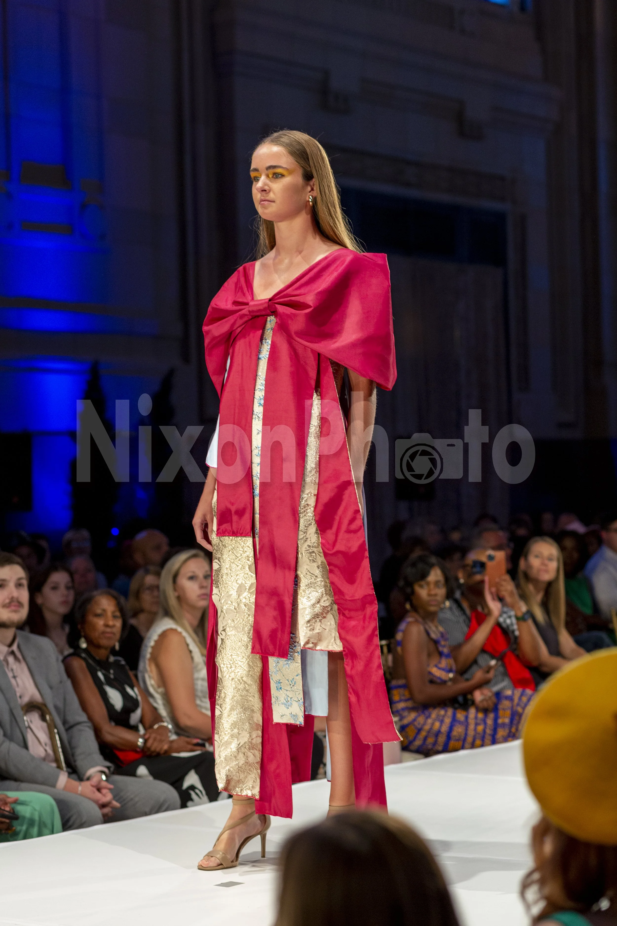Model on runway wearing a red bow dress with gold accents at a fashion show.