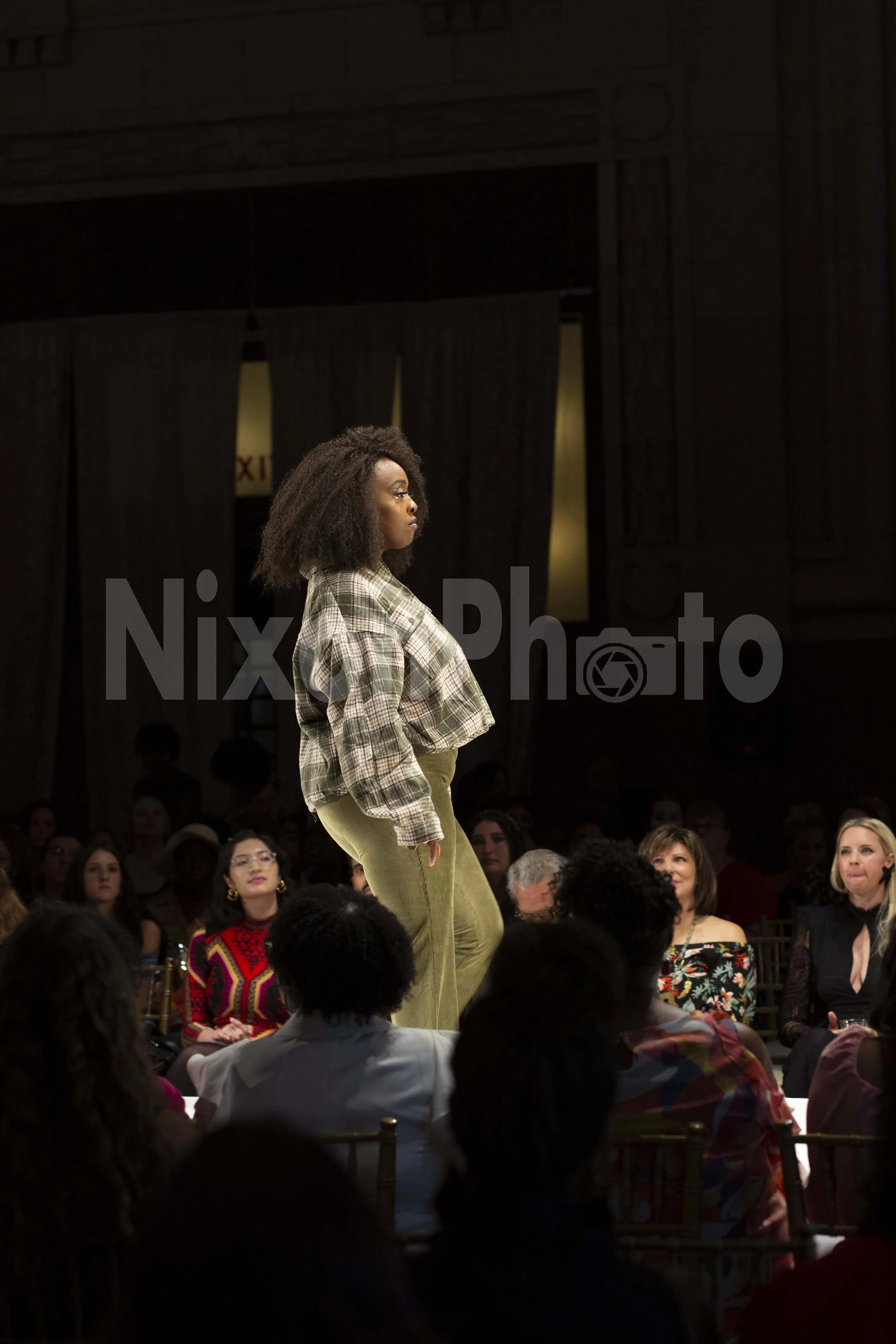 Model walking on a runway during a fashion show, wearing a plaid top and green pants, with an audience seated around the stage.