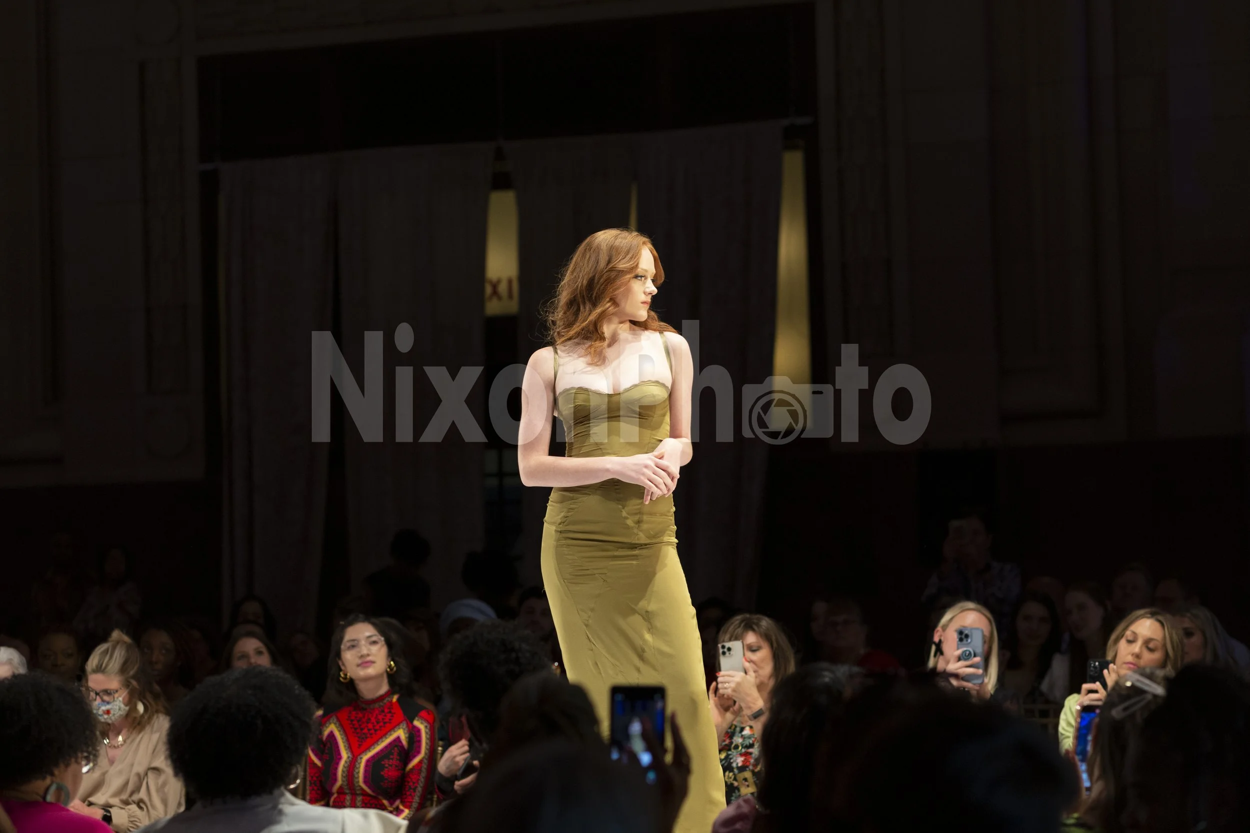 Model in yellow dress walking on a runway during a fashion show with audience taking photos.