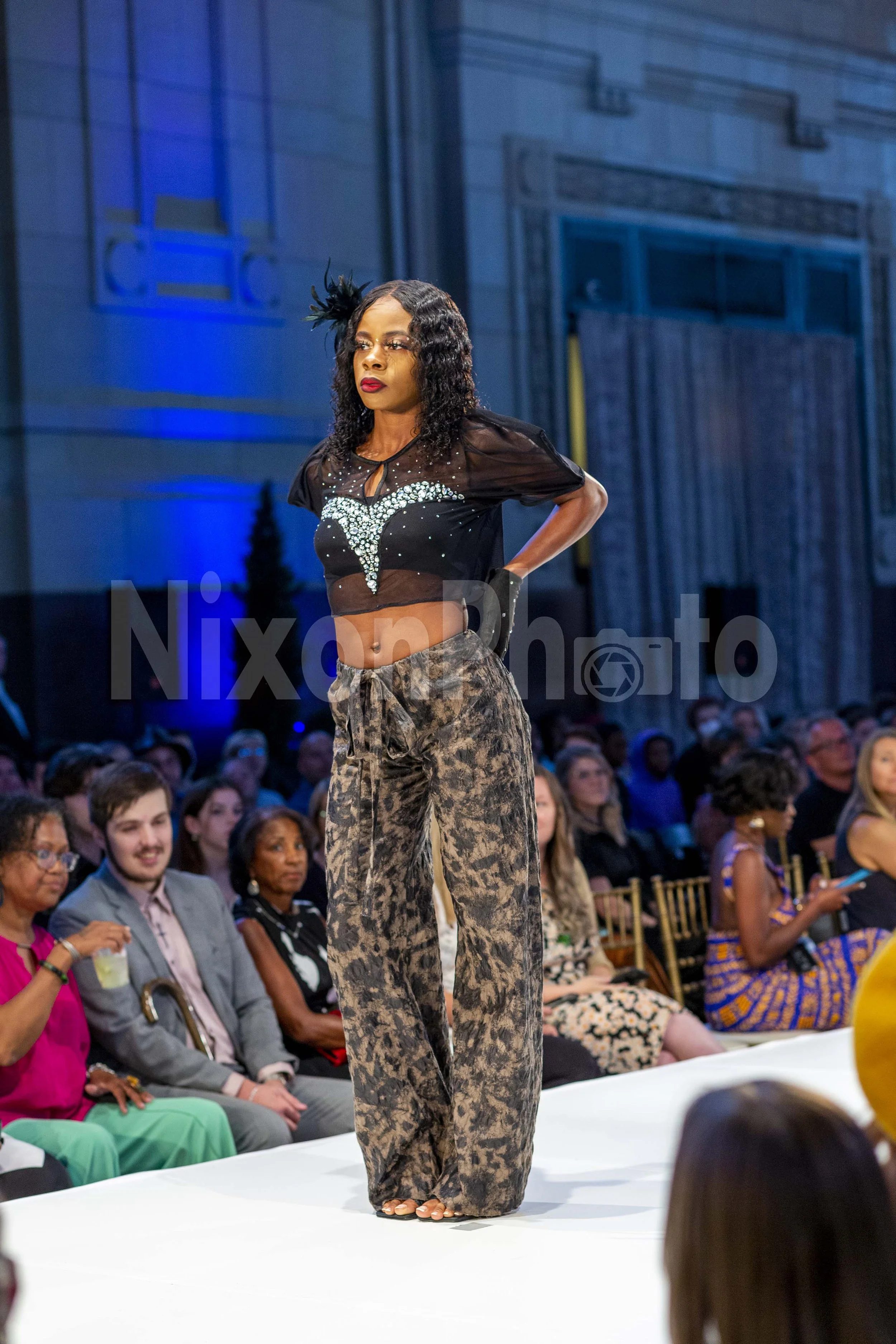 Woman modeling fashion on runway at event, wearing a black sheer top and patterned pants, audience in background.