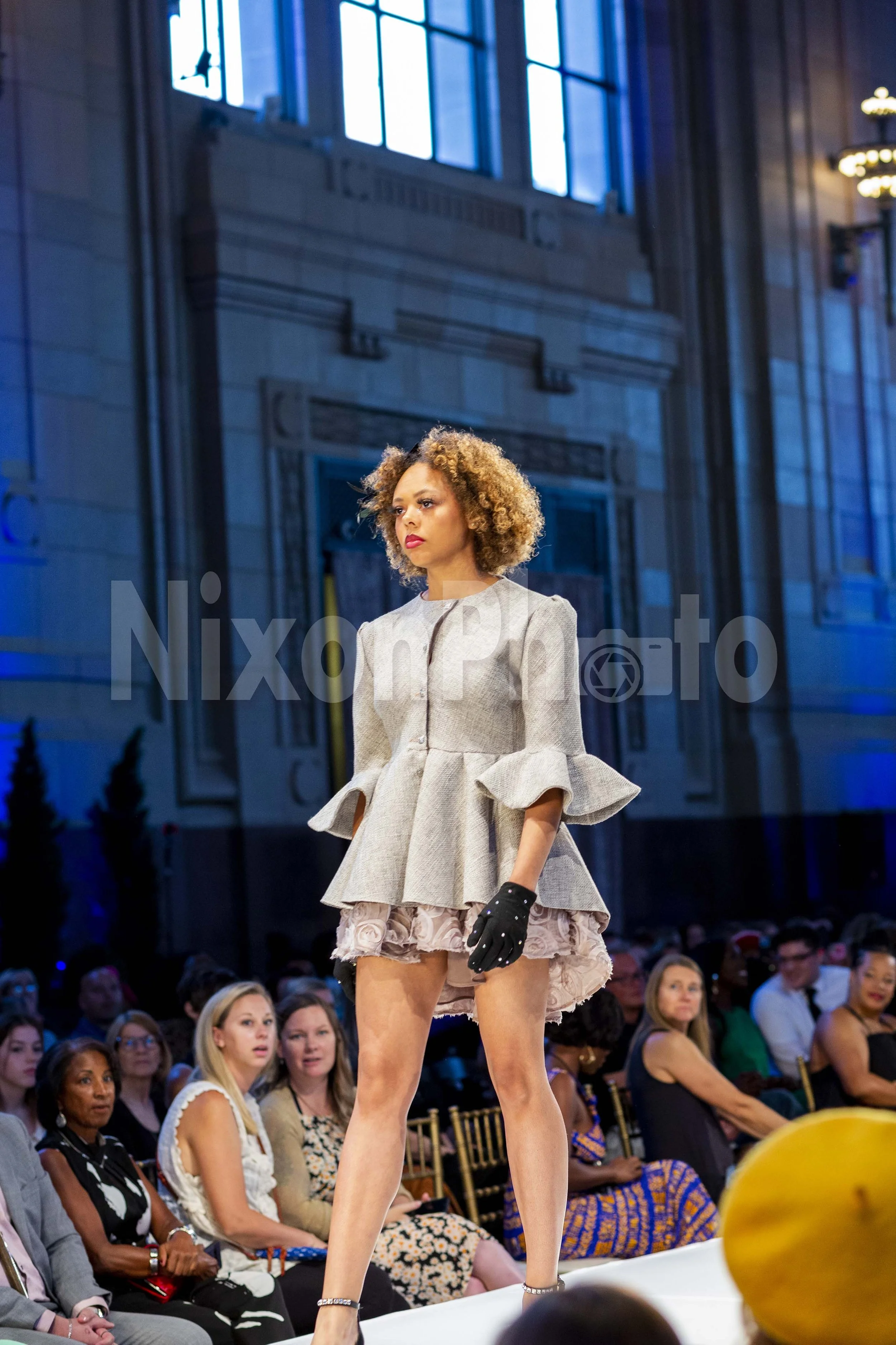 Model wearing gray blazer and lace skirt on runway at fashion show.
