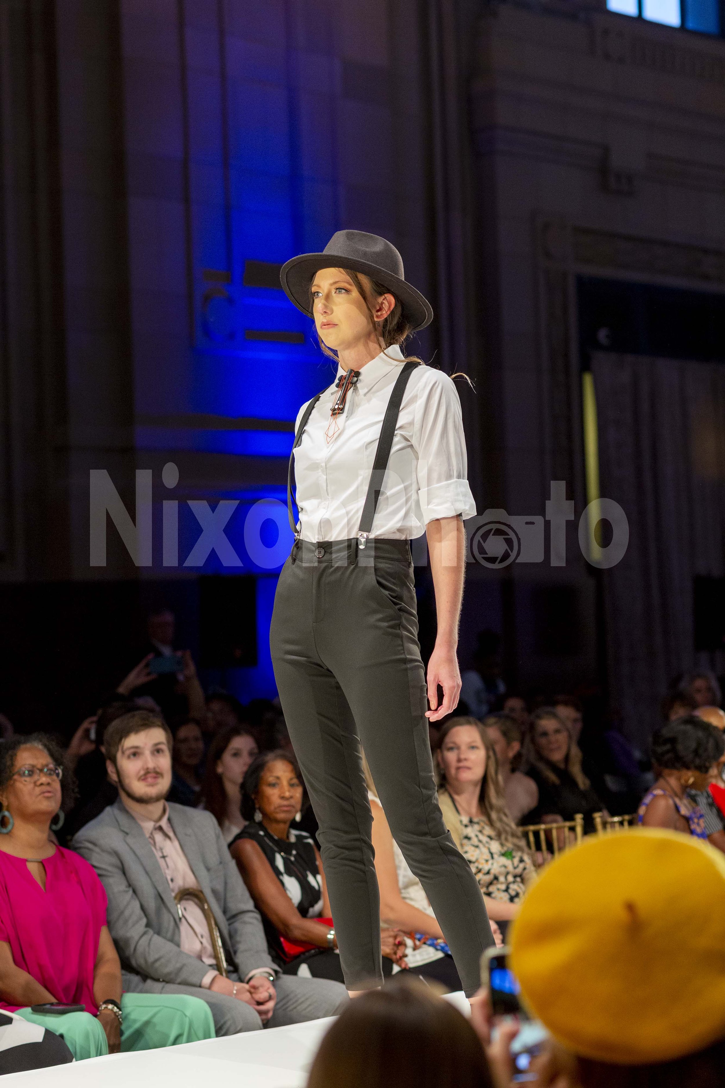 Model walking on the runway wearing a white shirt, suspenders, and hat in front of an audience.