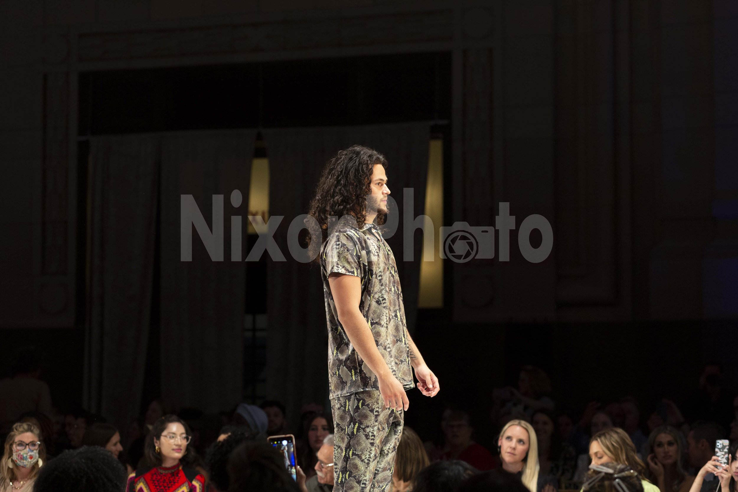 A man with long curly hair walks on a fashion runway wearing a snake print outfit, surrounded by seated audience members.