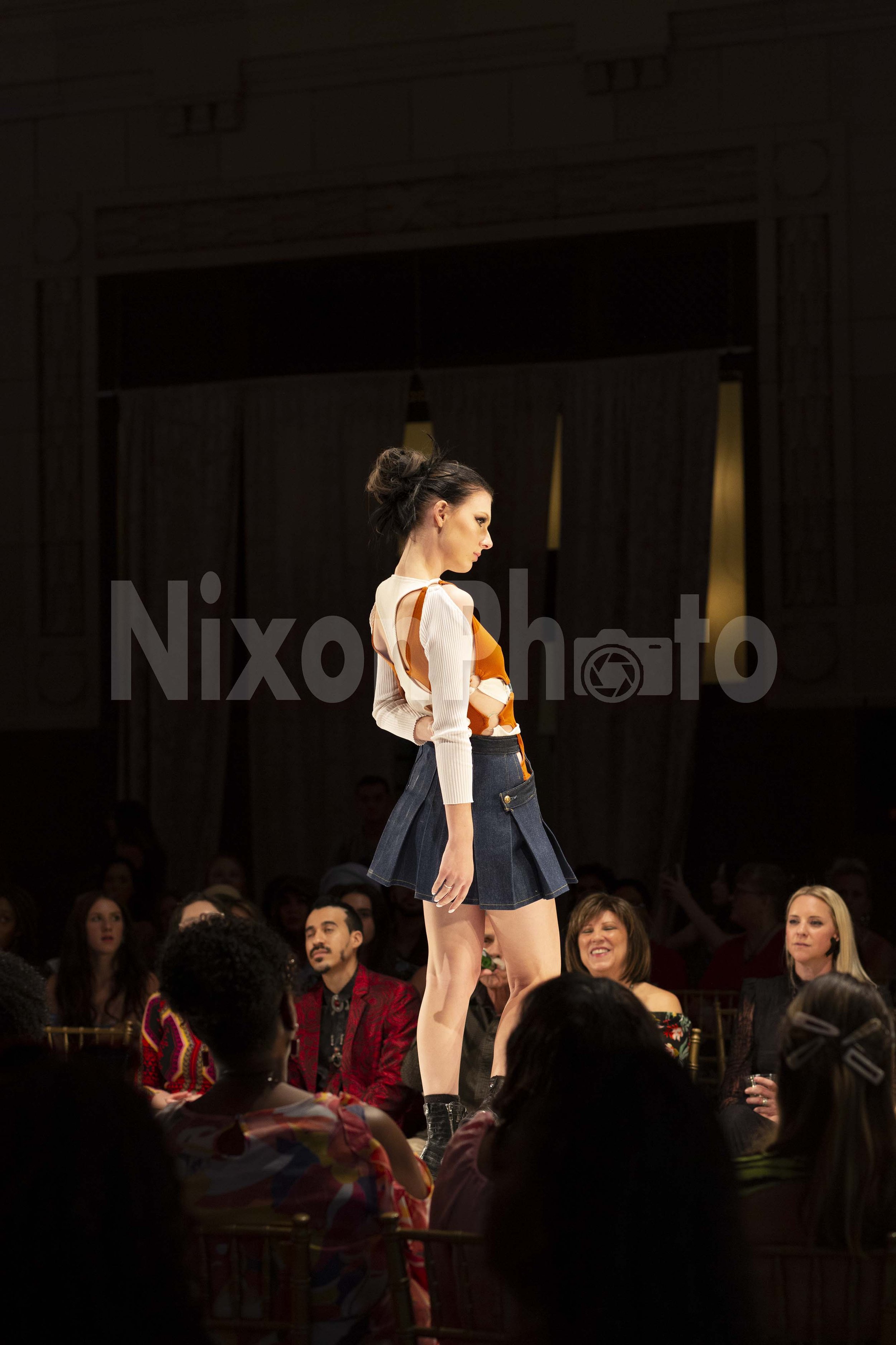 Fashion model walking on a runway during a fashion show, wearing a layered top and denim mini skirt with an audience observing.