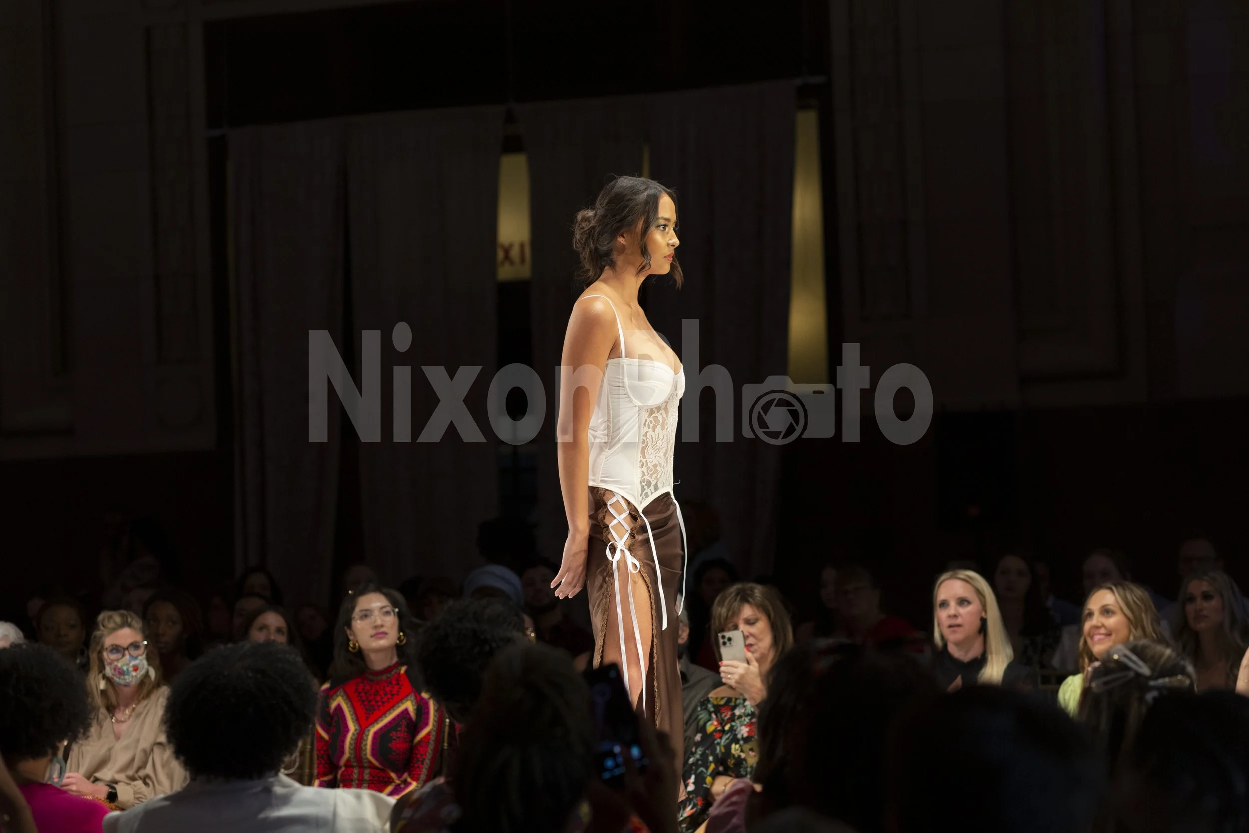 Model walking on a runway at a fashion show, wearing a white-patterned outfit. Audience members are seated on either side, some taking photos.