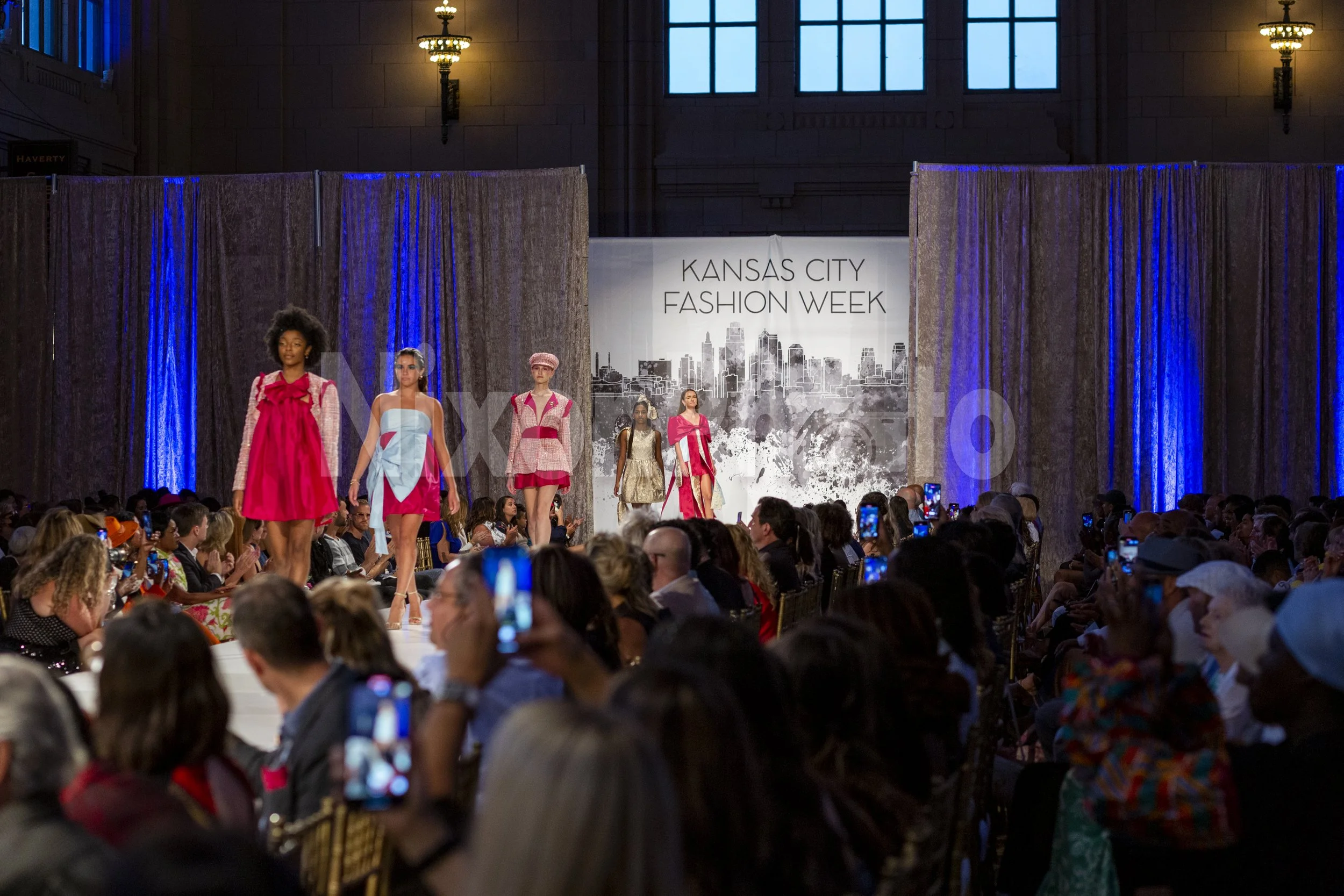 Models walking on a runway at Kansas City Fashion Week with an audience taking photos.