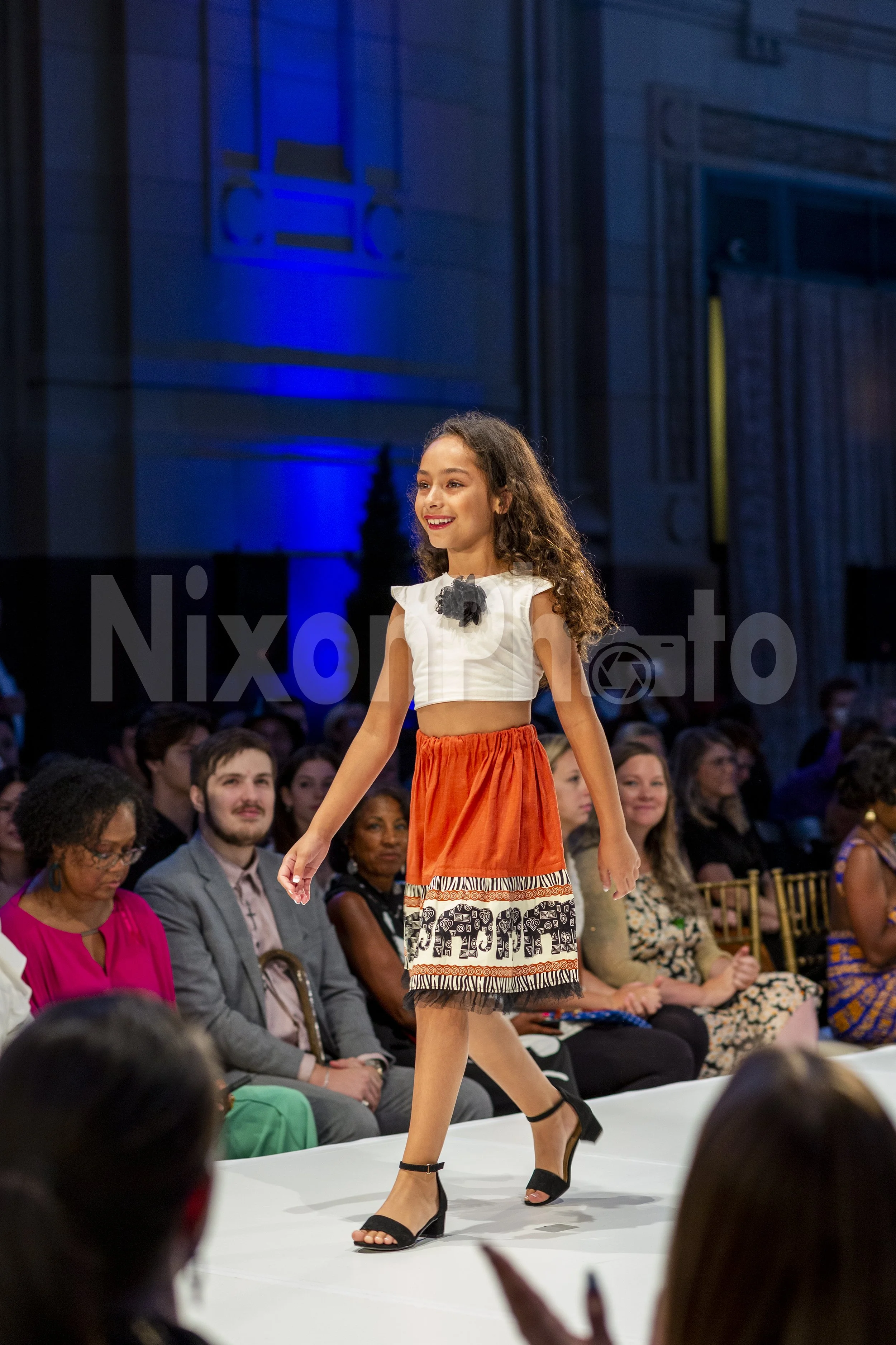 A young girl walking on a fashion runway wearing an orange and white outfit with elephant patterns. Audience members are seated around the runway watching the show. The background features blue lighting and architectural designs.