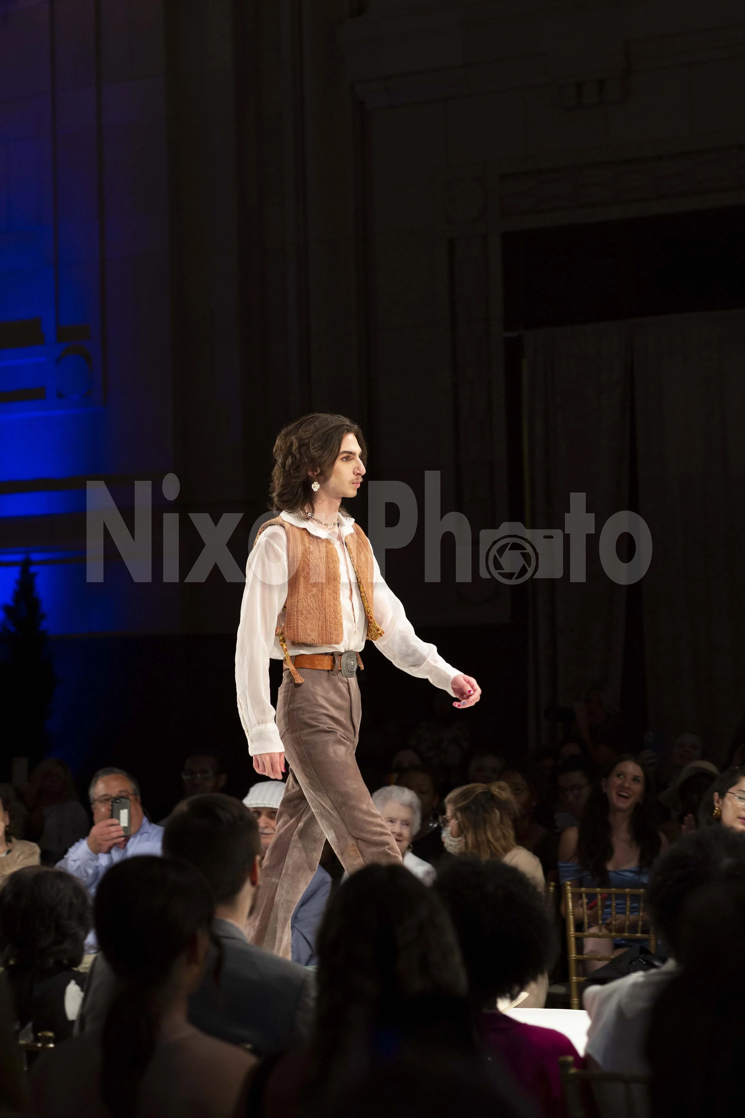 Model walking on a runway in brown vest and pants with an audience in the background.