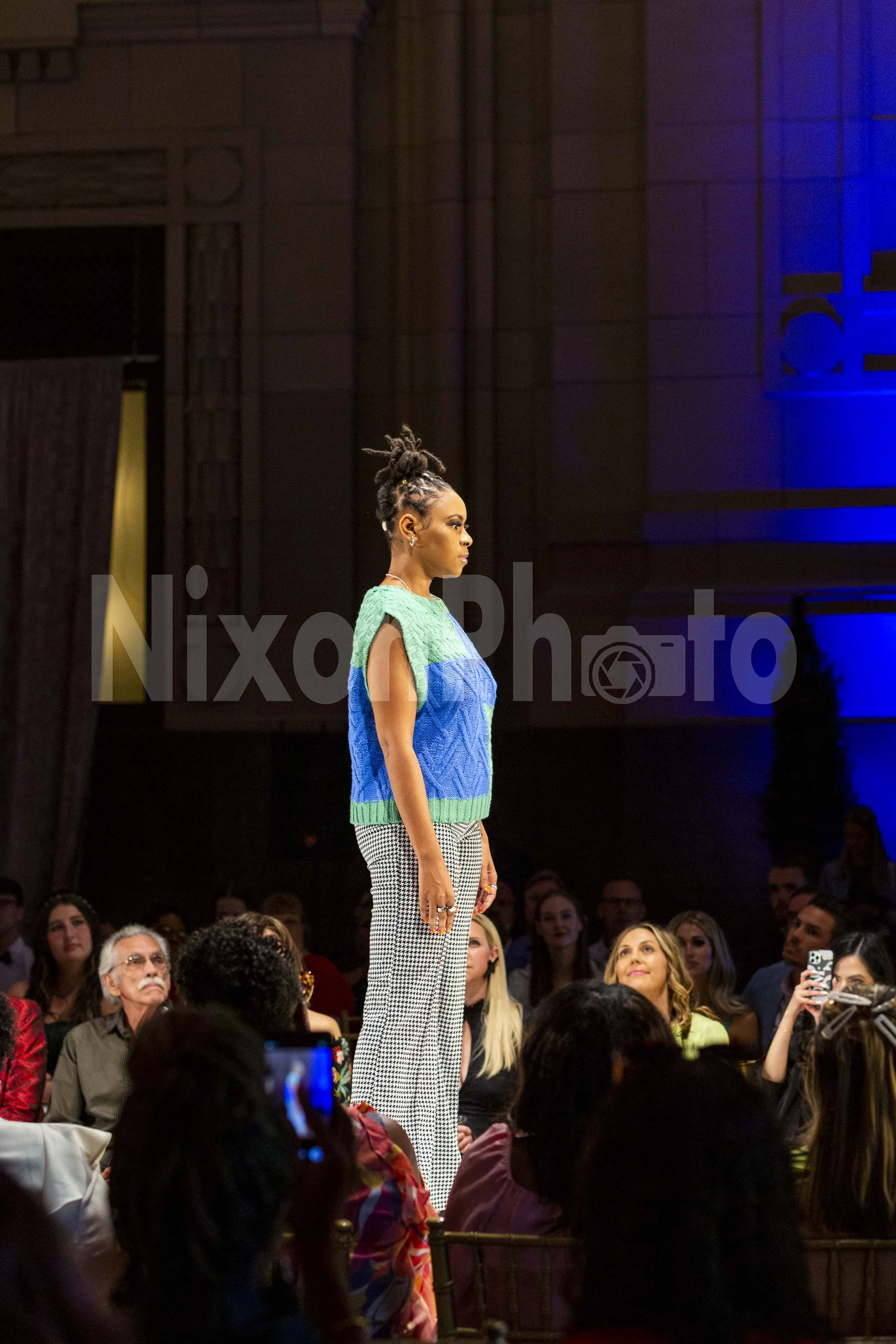 Model walking on a runway wearing a blue and green top with checkered pants, with an audience watching.