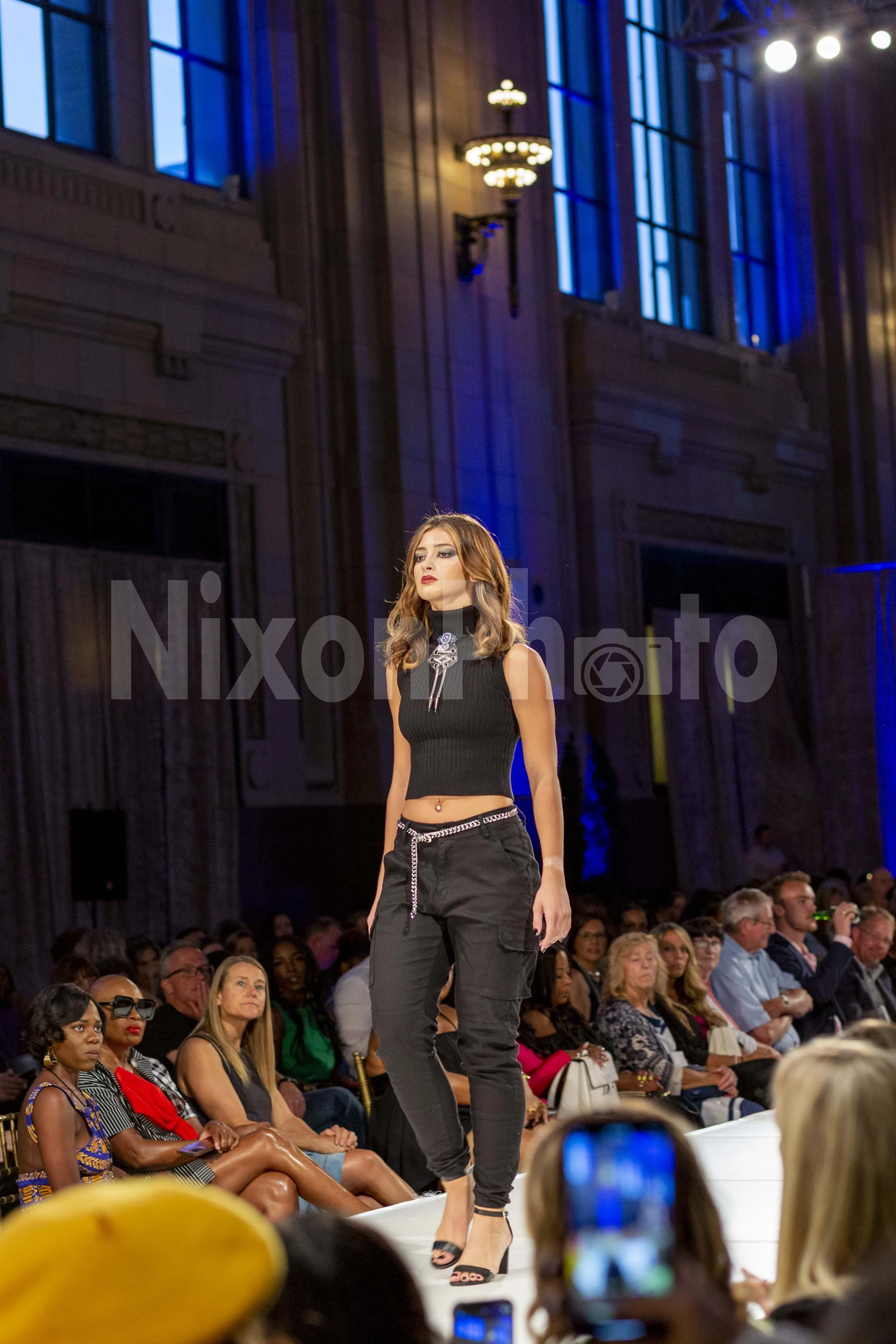 Model walking on a runway in a fashion show wearing a black top and pants, with an audience in the background.