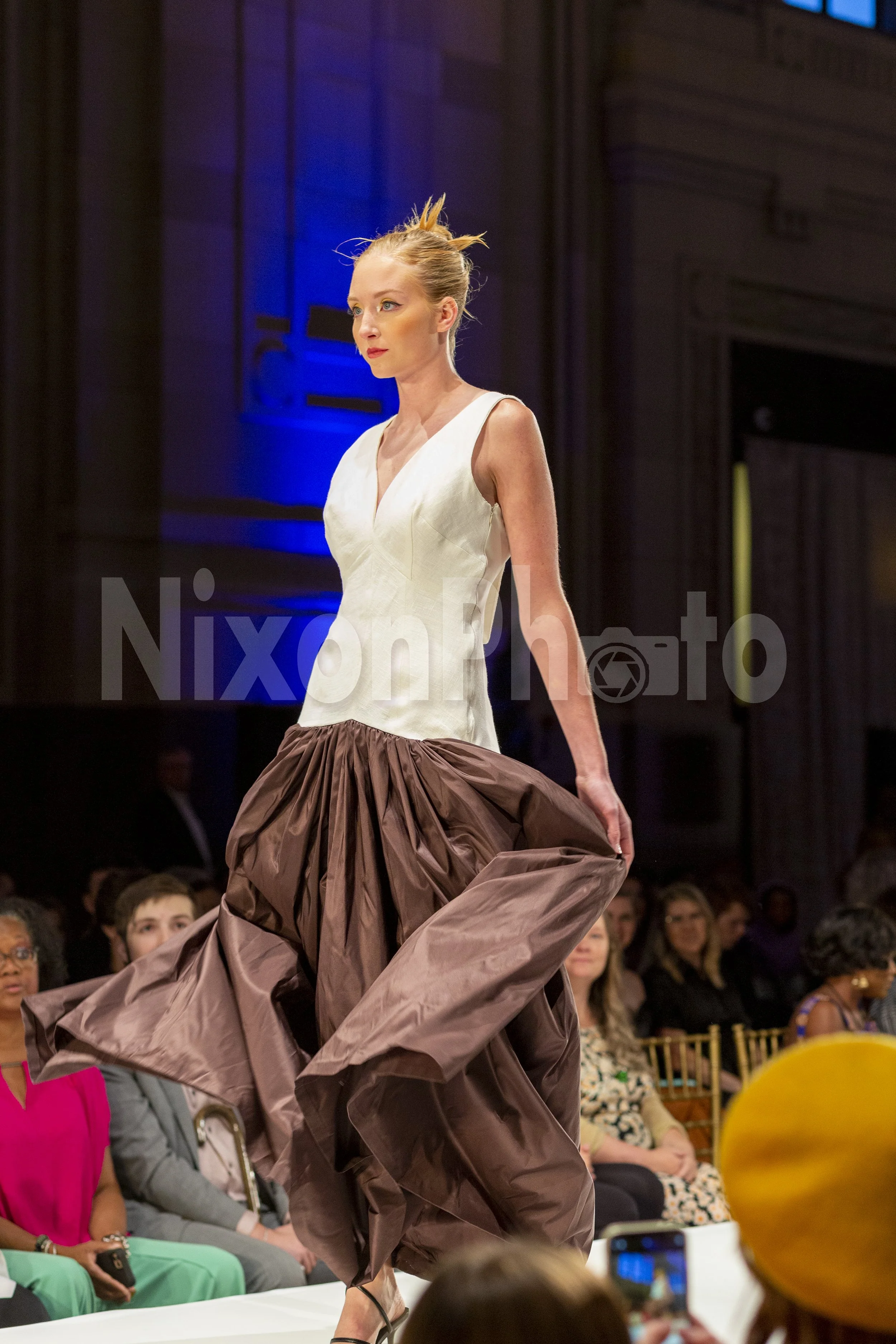Woman modeling a white top and voluminous brown skirt on a runway.