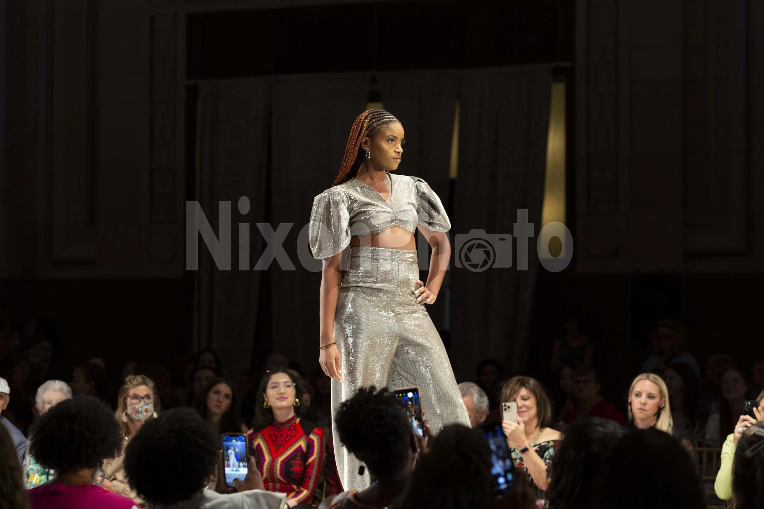 Model in metallic outfit on fashion runway with audience taking photos.