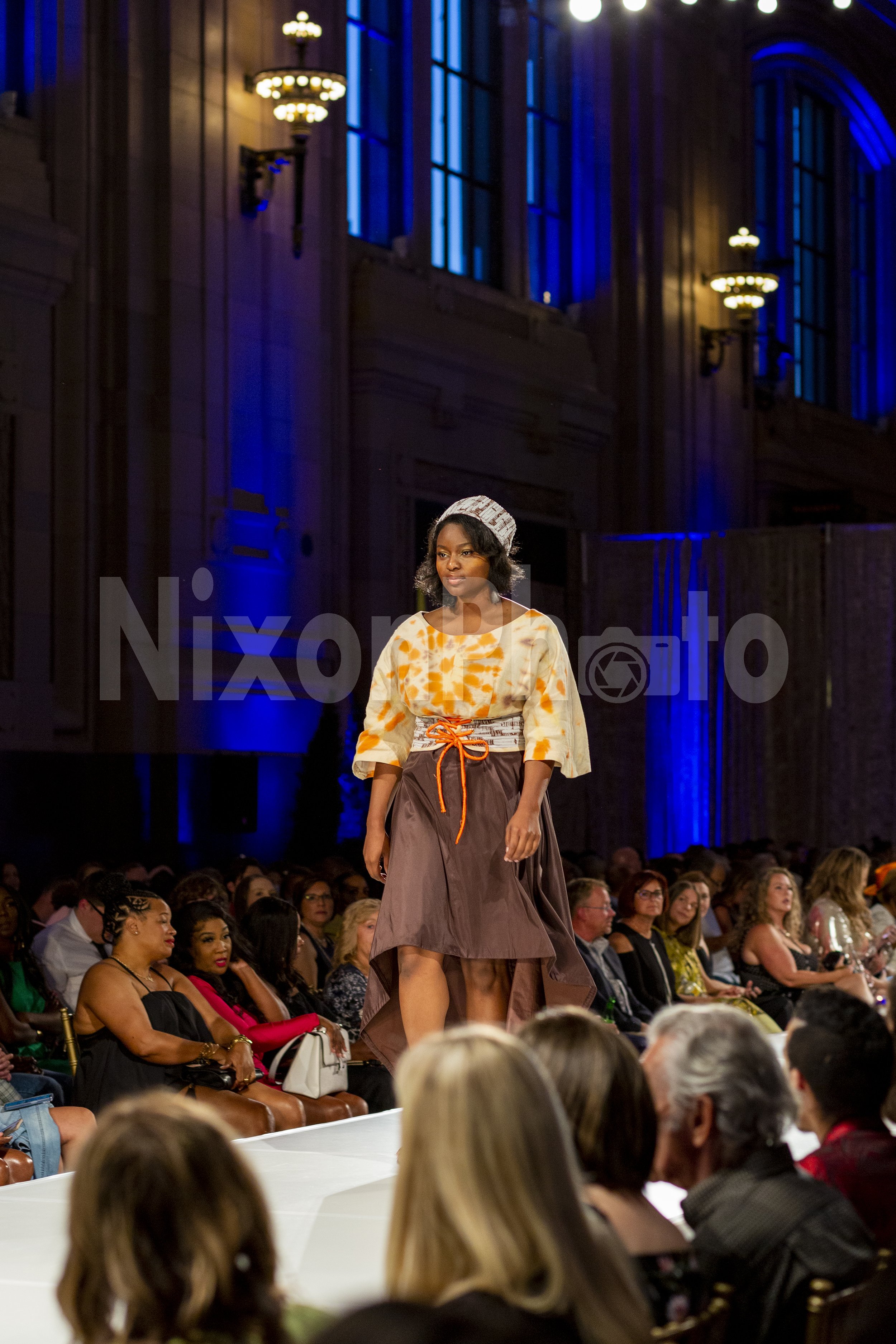 Model walking down a runway wearing a tie-dye blouse, brown skirt, and headscarf at a fashion show.