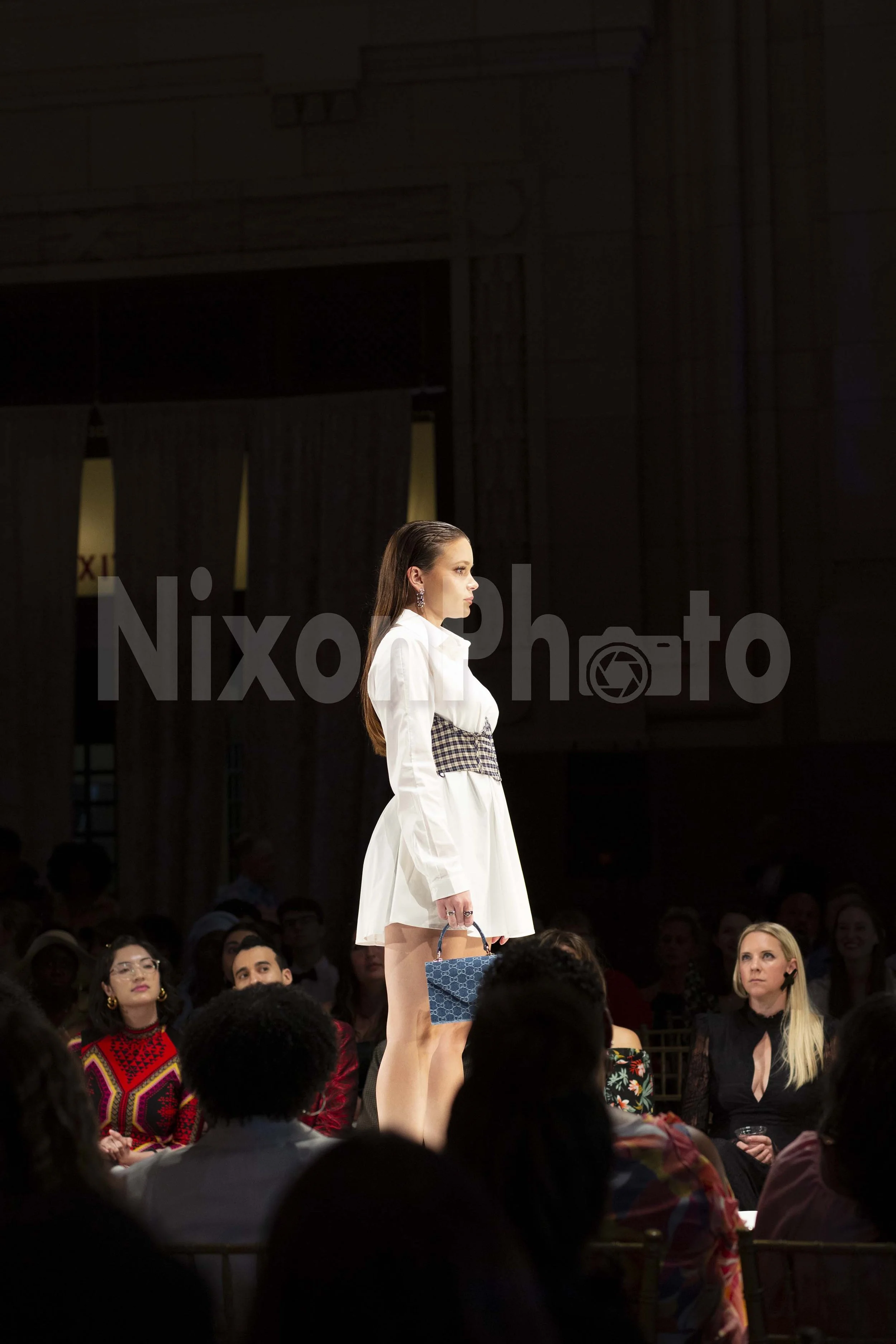 Model on a fashion runway wearing a white outfit with a checked corset, holding a small blue handbag. An audience is seated around the runway.