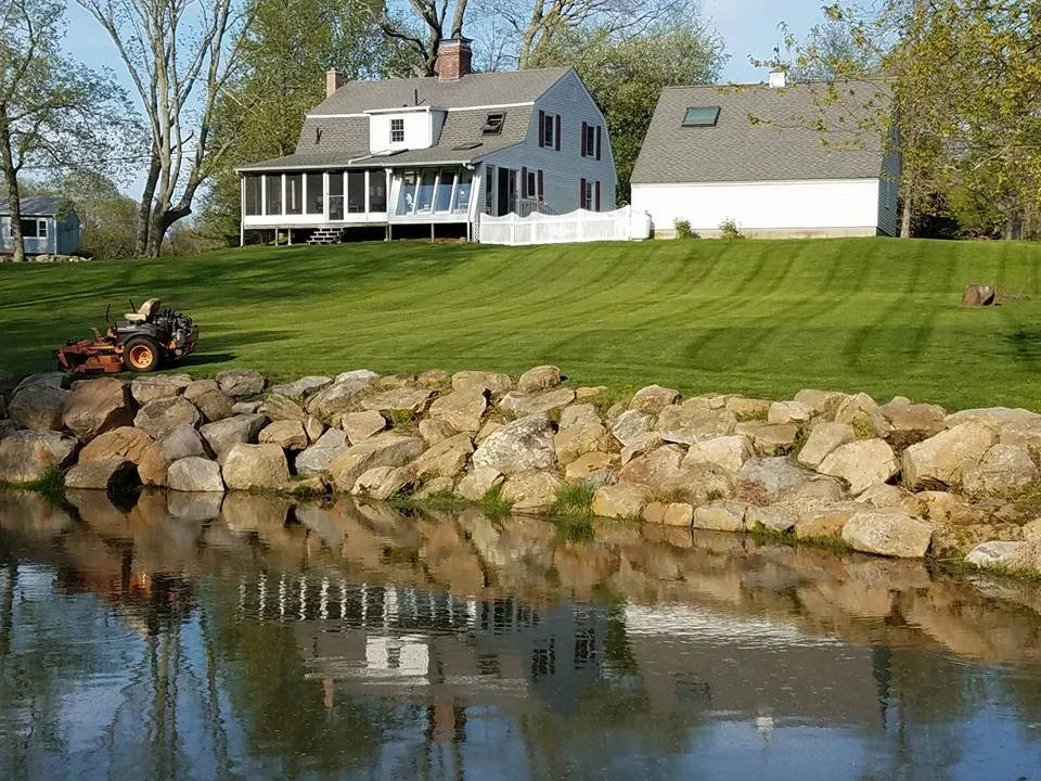 Freshly mowed lawn above a stone-edged pond with a white farmhouse behind