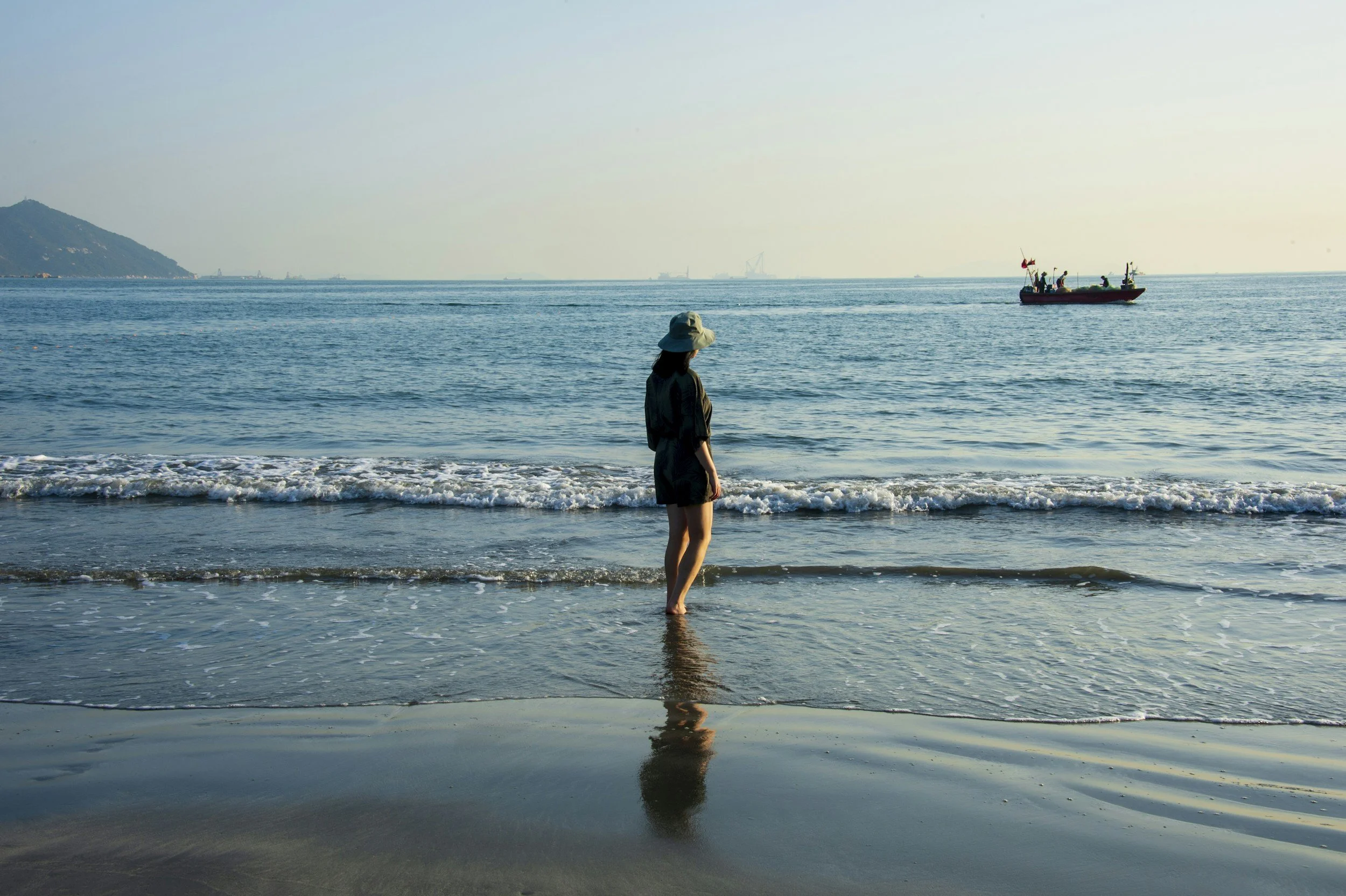 A woman standing on the beach near the water, wearing a large sun hat and dark clothing, with a boat sailing in the distance on the calm ocean under a clear sky.