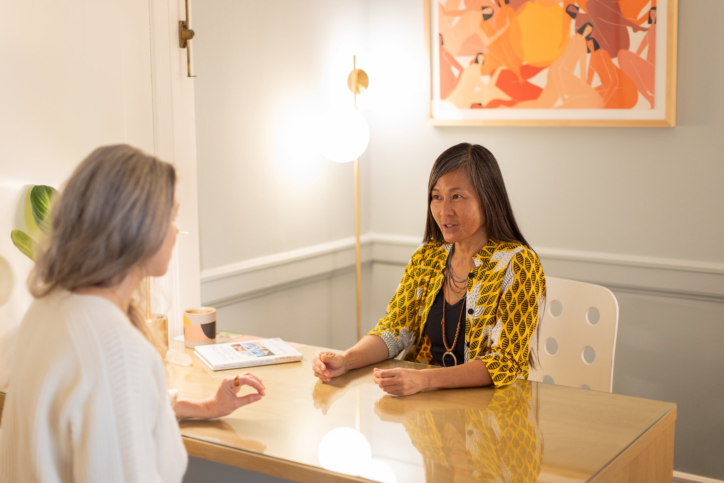 Michelle Kuroda, LAc, master acupuncturist in San Francisco, CA and Kailua-Kona, HI, specializing in nervous system regulation, neuromuscular re-education, embodied healing, and energy alignment speaking with a client in her office.