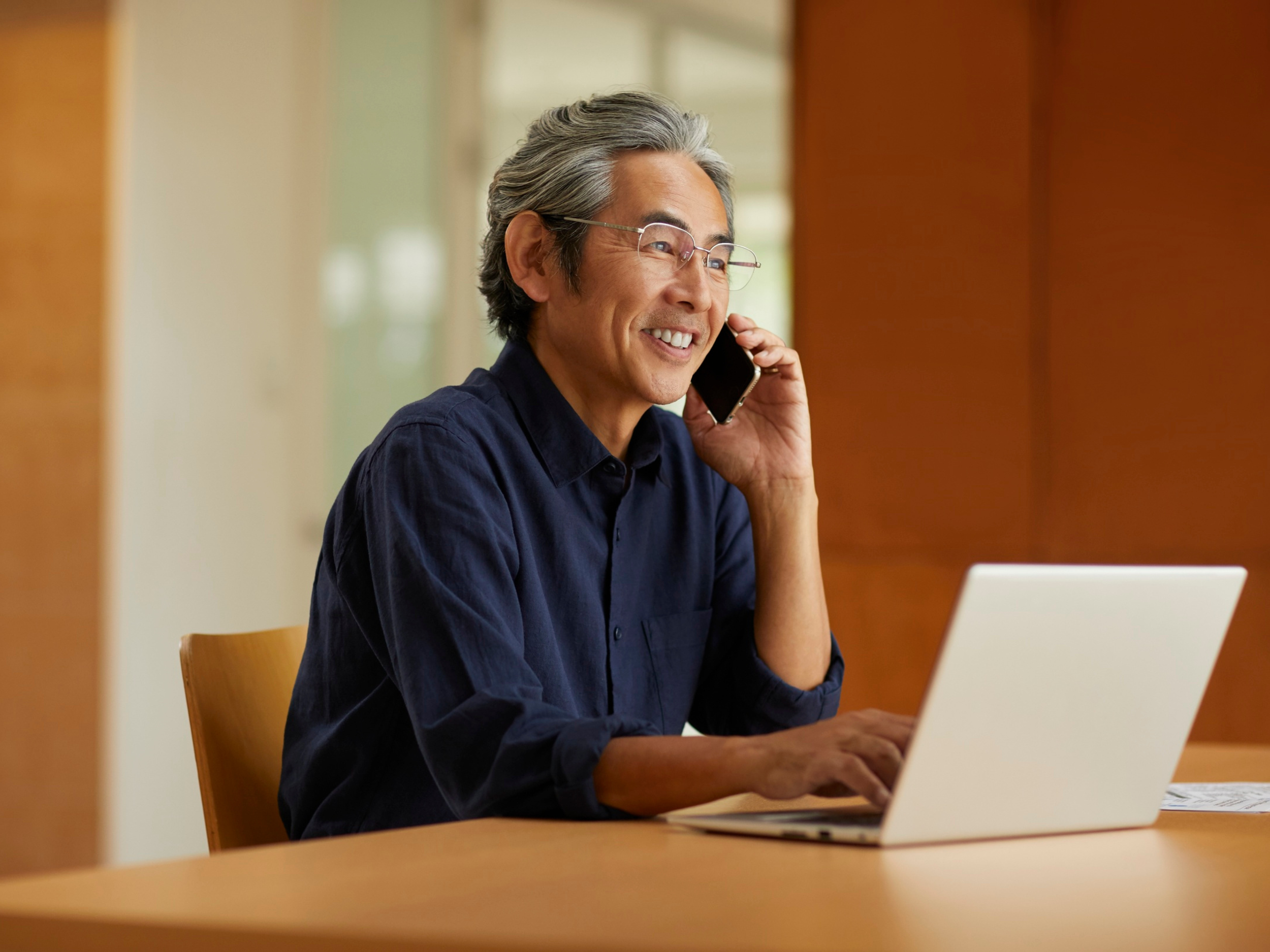 A woman with curly brown hair, wearing a light blue striped shirt, sitting on a beige sofa, smiling and looking at a laptop in front of her.