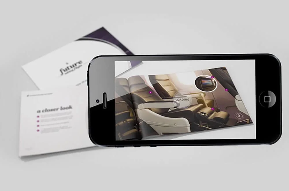 Smartphone capturing an augmented reality view of airplane seats with pink markers on a printed brochure and a laptop in the background.