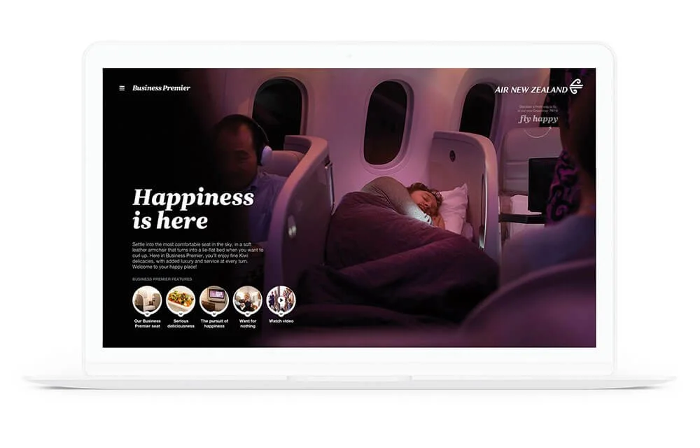 A laptop screen displaying a webpage with a woman sleeping on an airplane seat, passenger and window behind her, text saying 'Happiness is here' and airline branding 'AIR NEW ZEALAND'.