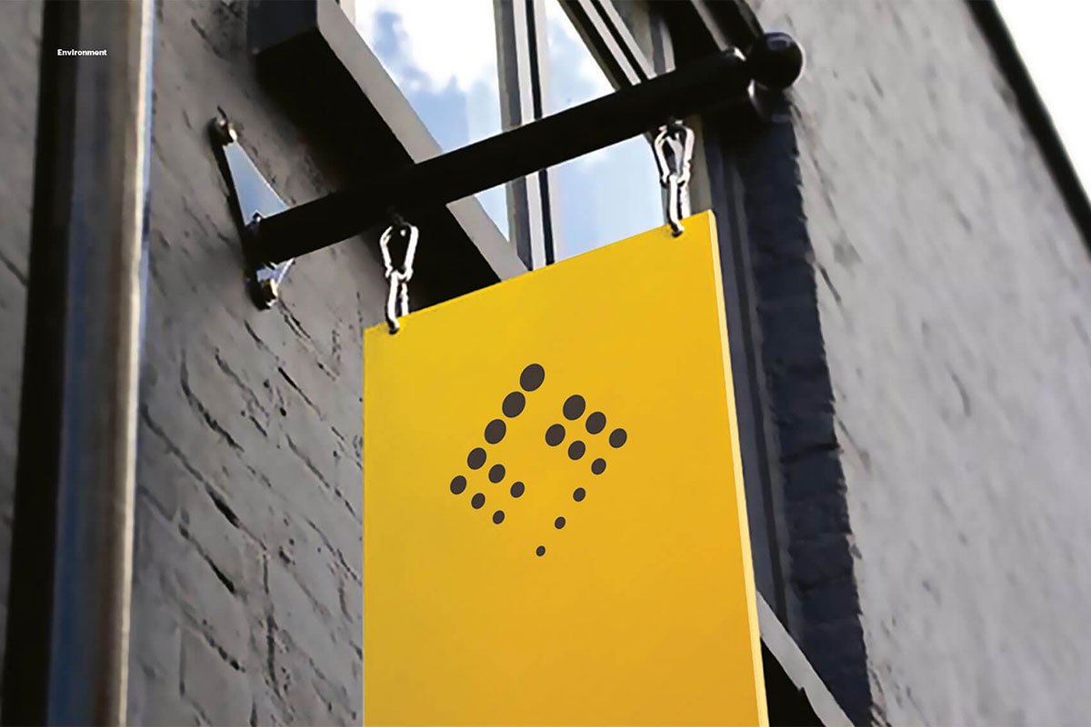 Kevin Schluter Design | Shimmer Branding | Application | A yellow sign with black braille dots hanging from a black metal bar outside a building with white brick walls.