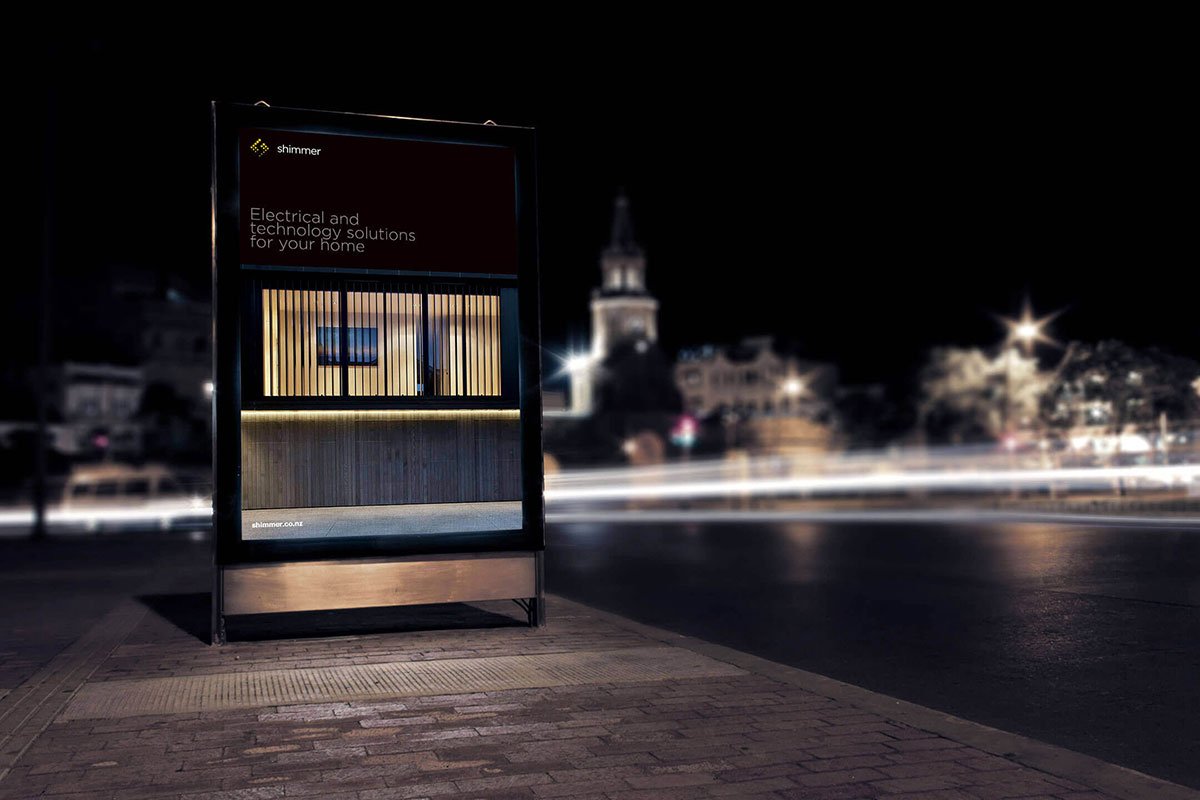 Kevin Schluter Design | Shimmer Branding | Application | Bus stop shelter with advertisement at night with blurred cityscape and streetlights in the background.