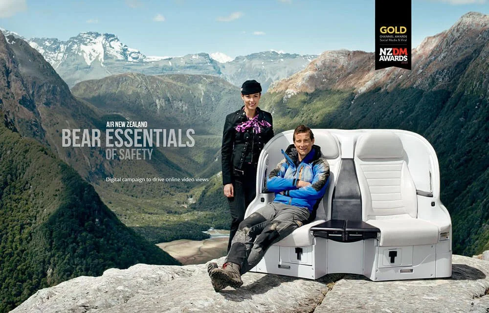 Two people, a woman and a man, are posing outdoors on a large rock with mountains and a river in the background. The woman is standing, and the man is sitting on a white mountain safety seat. There are logos and text on the image, including 'Air New Zealand', 'Bear Essentials of Safety', and awards logos.
