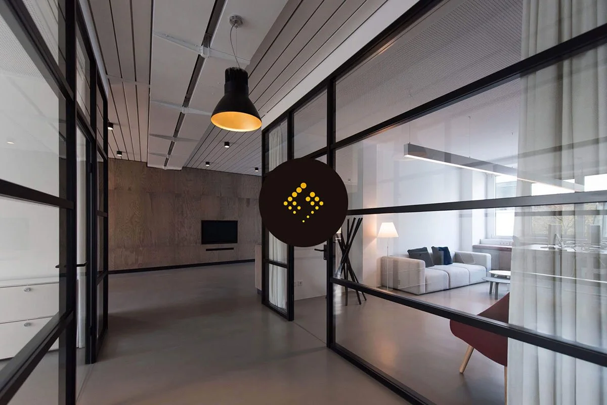 Kevin Schluter Design | Shimmer Branding | Application | Modern interior with glass partitions, a hanging black and yellow lamp, a white sofa, lamp, and window in the living area