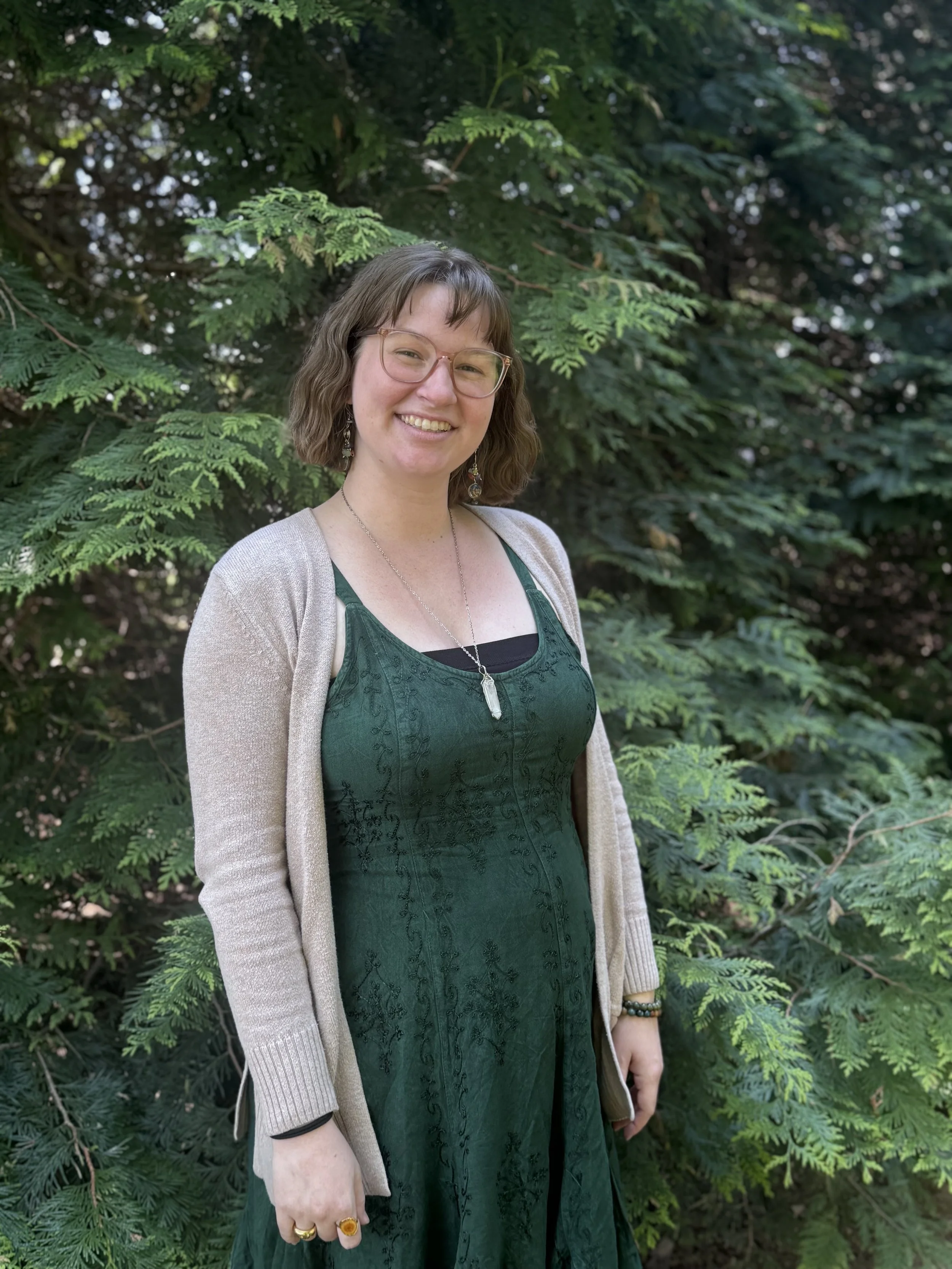 A woman with shoulder-length brown hair and glasses smiling, wearing a dark green dress and beige cardigan, standing outdoors in front of a lush green pine tree.
