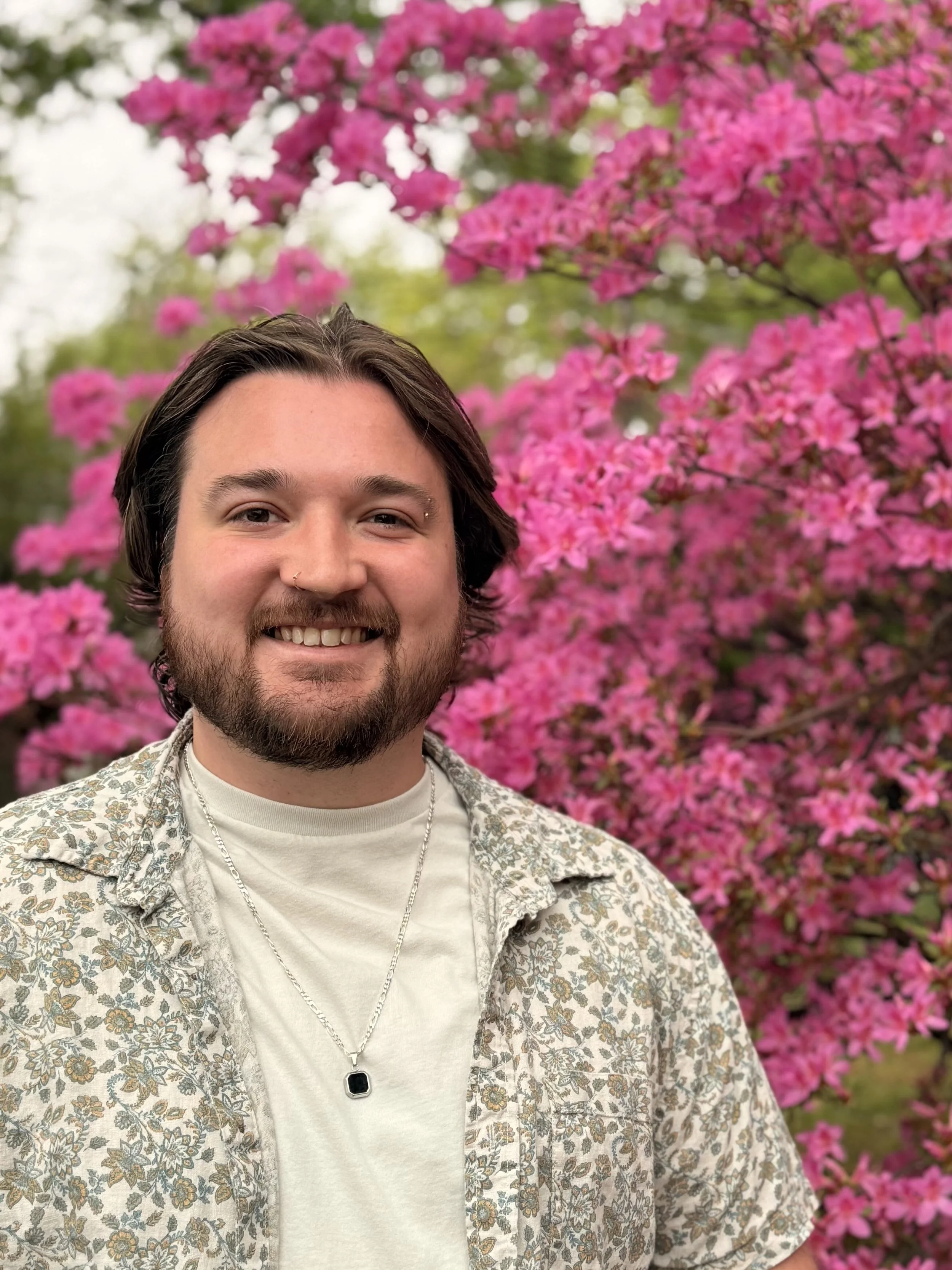 Close-up of a smiling young man with a beard, wearing a dark suit, white shirt, and a colorful patterned tie, outdoors with blurred green foliage in the background.