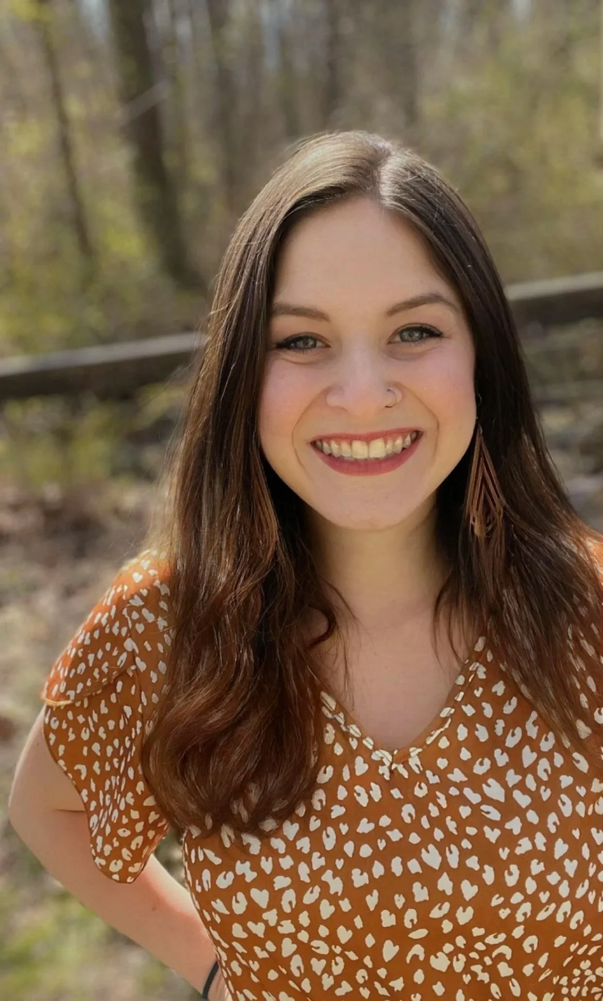 A smiling woman with long, wavy brown hair, wearing a rust-colored dress with white heart-shaped patterns and a nose ring, standing outdoors in a wooded area.