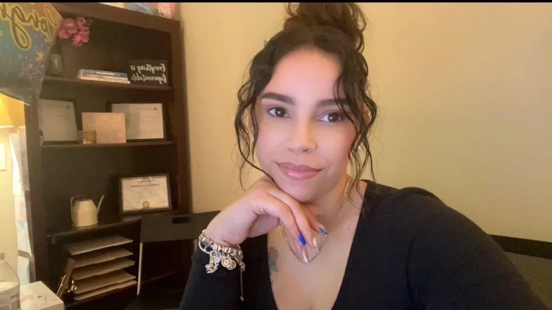 A woman with curly dark hair in a bun, wearing a black top, sitting indoors. She's resting her chin on her right hand, with colorful jewelry on her wrist, and has a slight smile. Behind her is a wooden bookshelf with framed certificates, books, and decorative items.