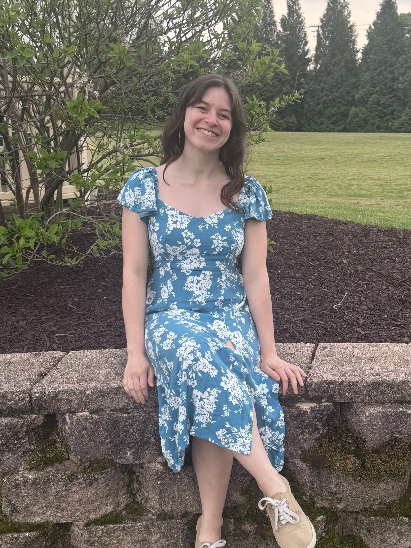 A young woman with brown hair wearing a blue and white floral dress, sitting on a stone ledge outdoors, smiling, with green shrubs, trees, and a lawn in the background.