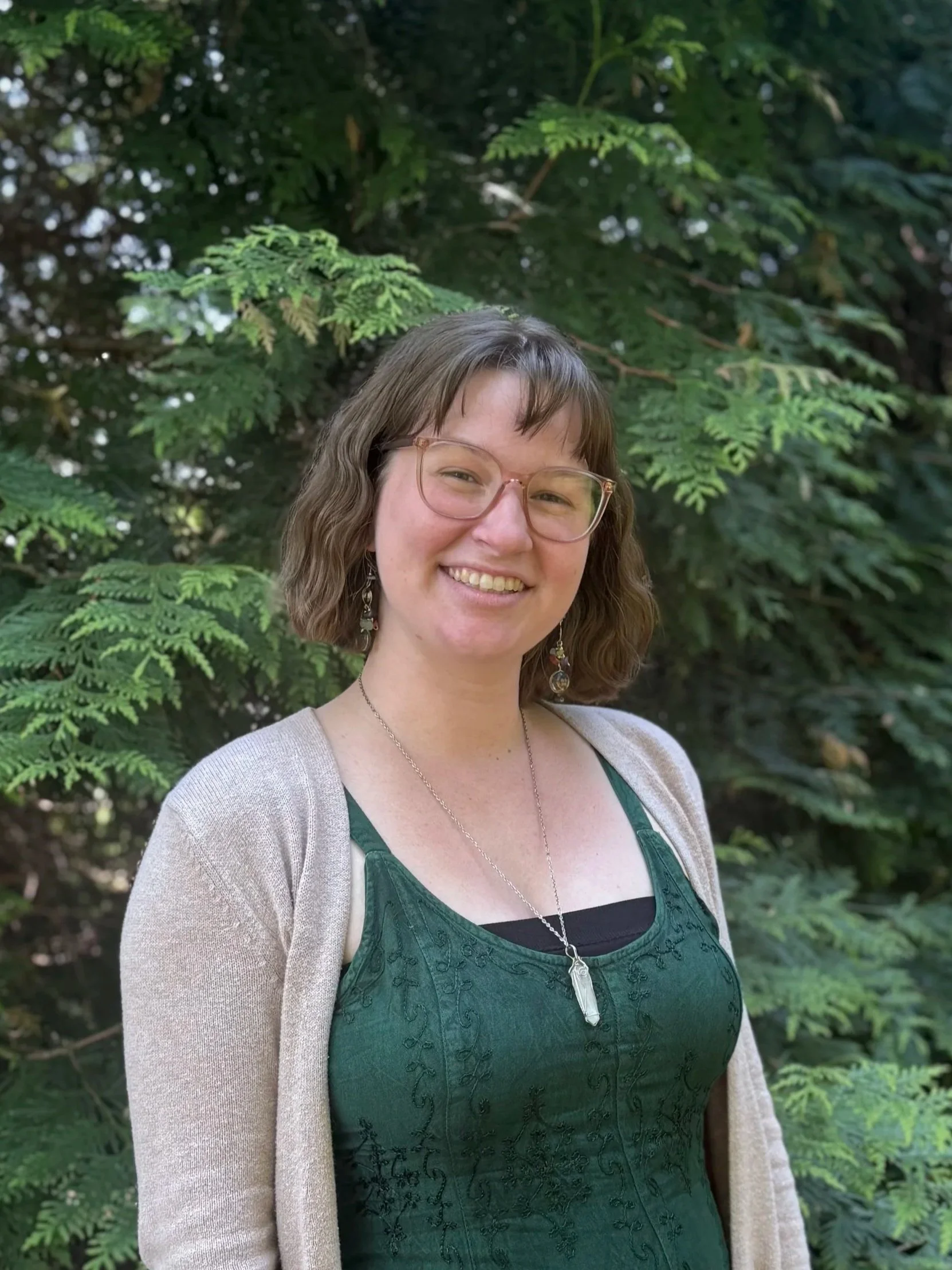 A woman with glasses and earrings smiling outdoors in front of a green leafy background.