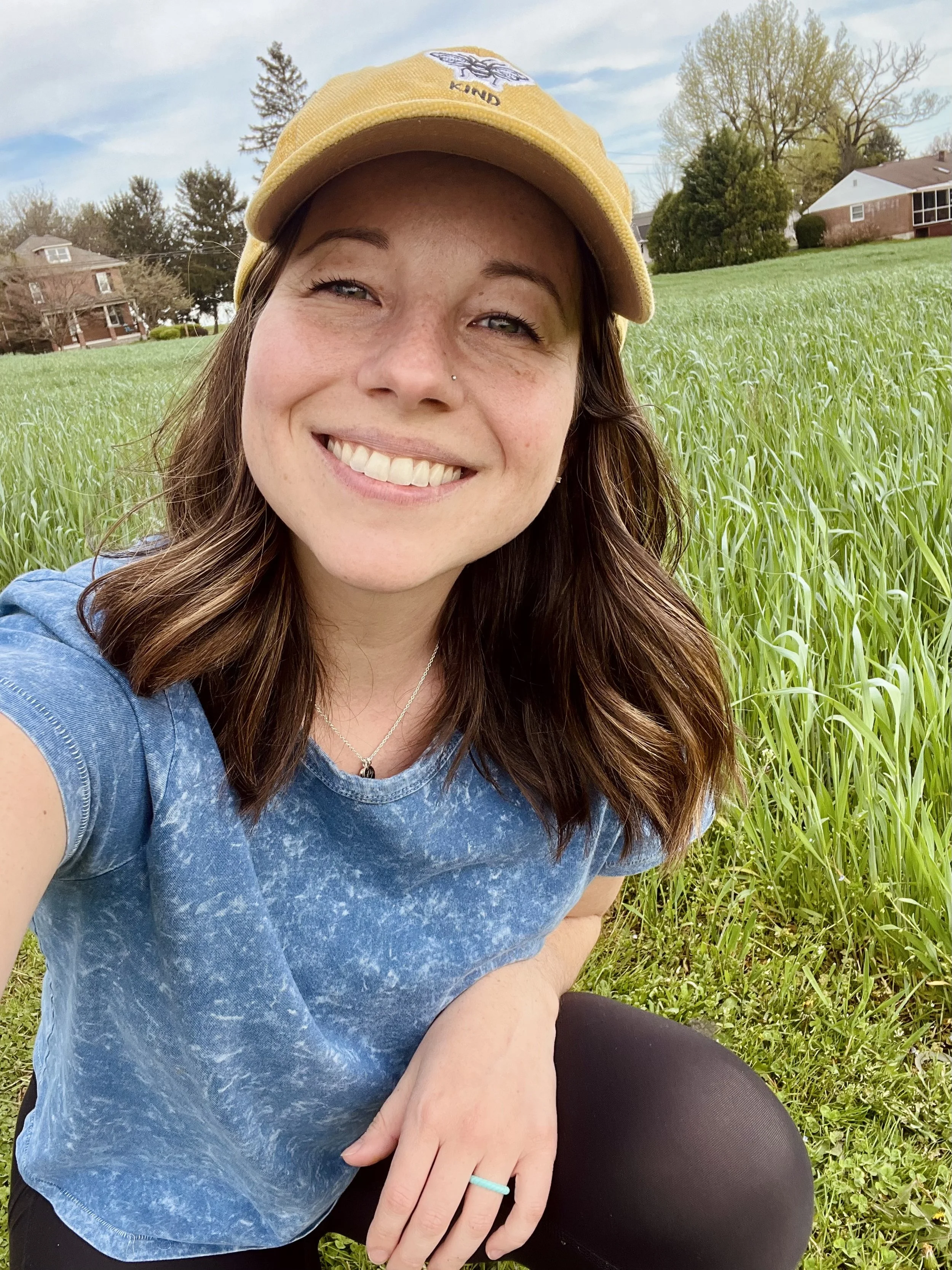 Close-up of a smiling woman with brown hair and light skin, wearing a yellow cap and a blue T-shirt, sitting on grass in a park with houses and trees in the background.
