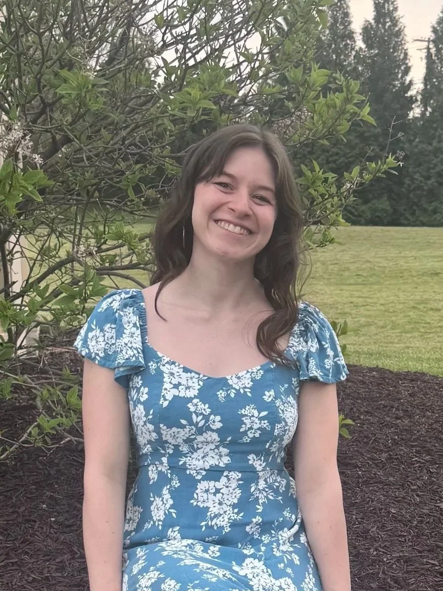 A young woman with long brown hair, wearing a blue and white floral dress, sitting outdoors on dark mulch, smiling, with green trees and grass in the background.