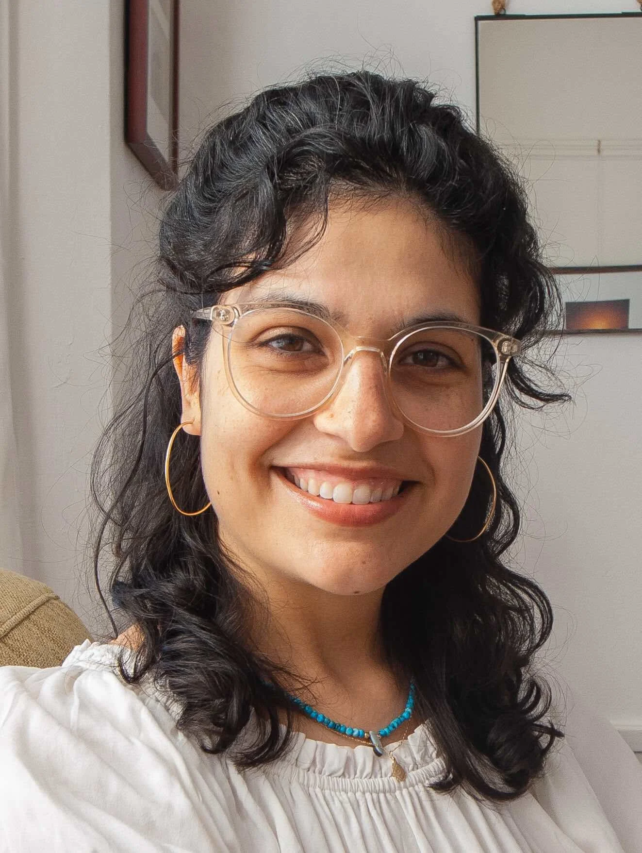Bay Area women’s therapist with dark curly hair wearing glasses, hoop earrings, a turquoise beaded necklace, and a white top, smiling indoors.