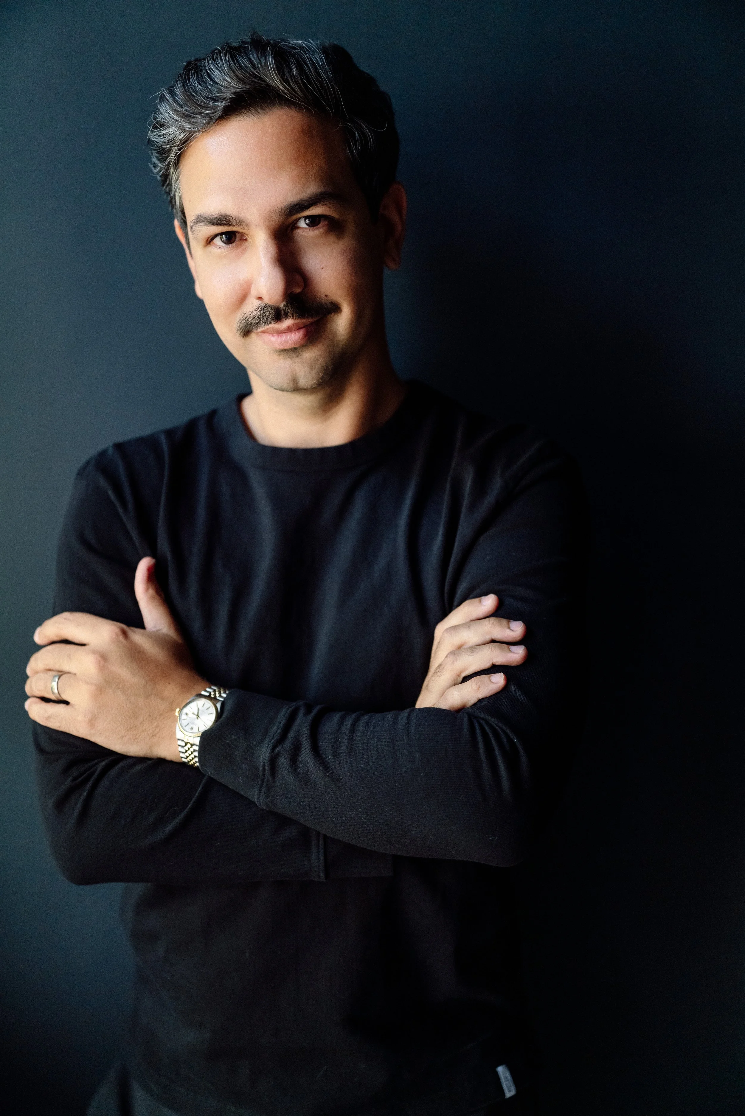 A man with dark hair and a mustache wearing a black long-sleeve shirt and a silver wristwatch, standing with arms crossed against a dark background.