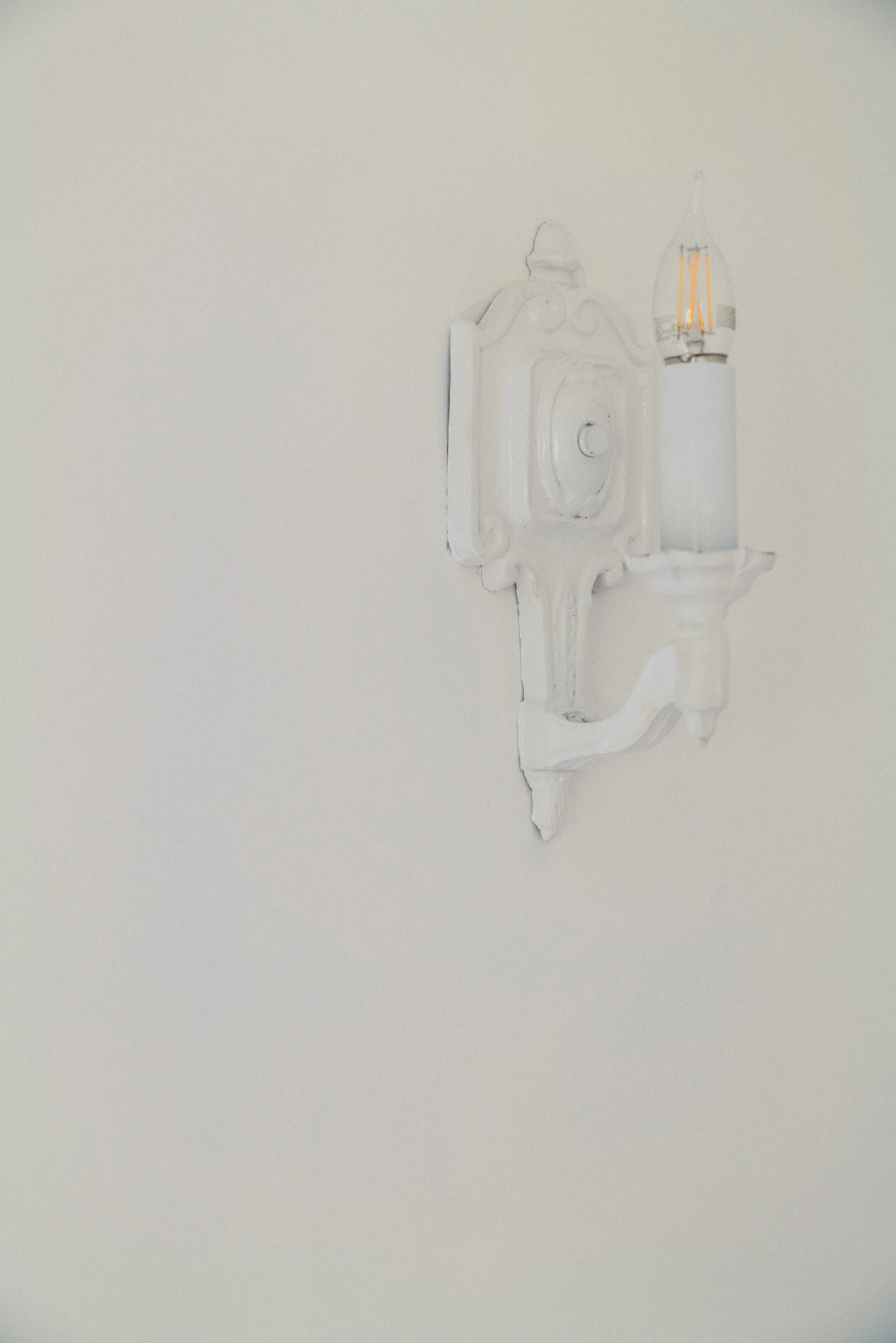 A vintage white wall-mounted lamp with an exposed lightbulb on a plain white wall.
