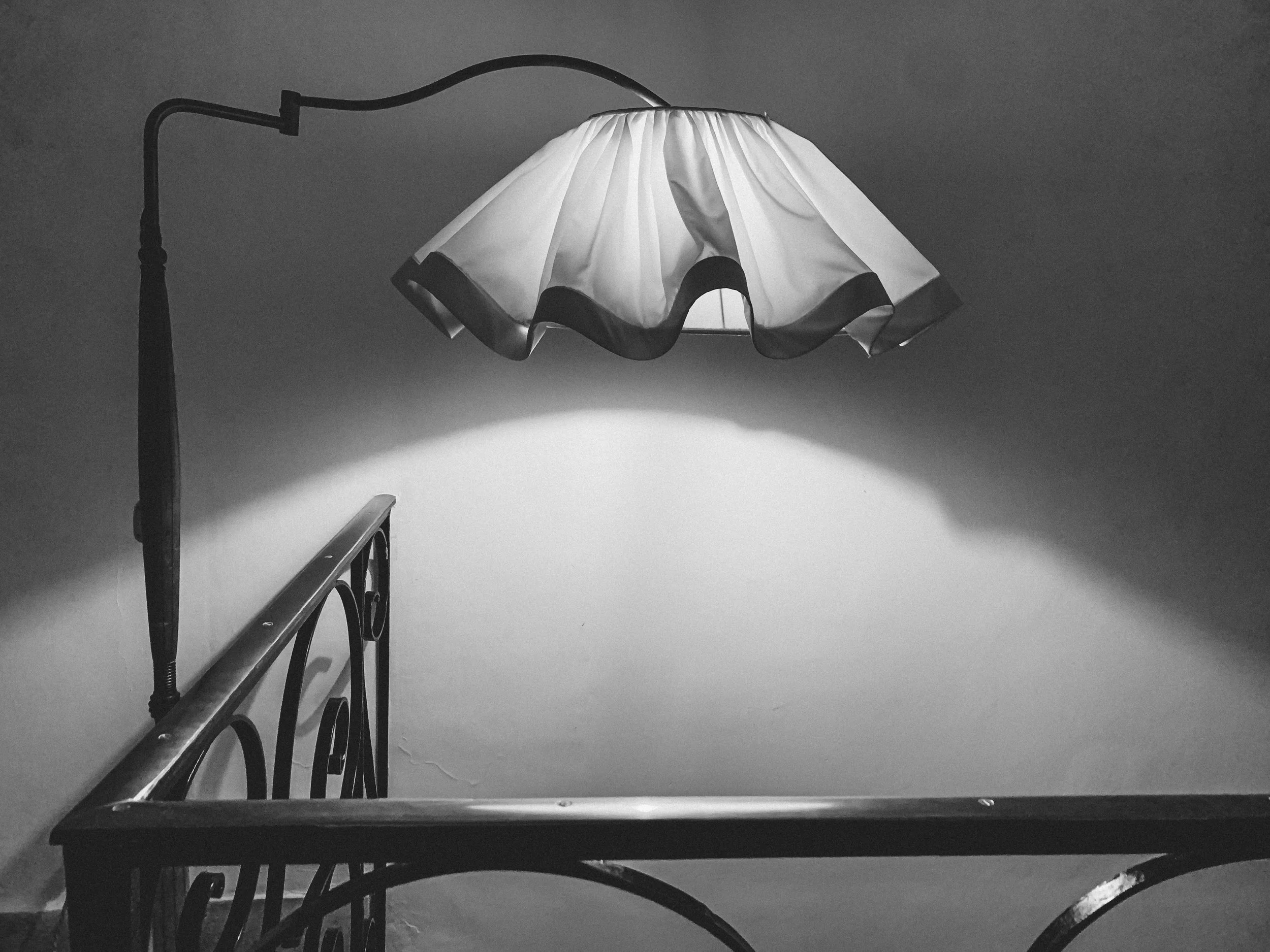 Black and white photo of a table lamp with a ruffled lampshade, casting light on a surface below, with a staircase railing visible in the foreground.