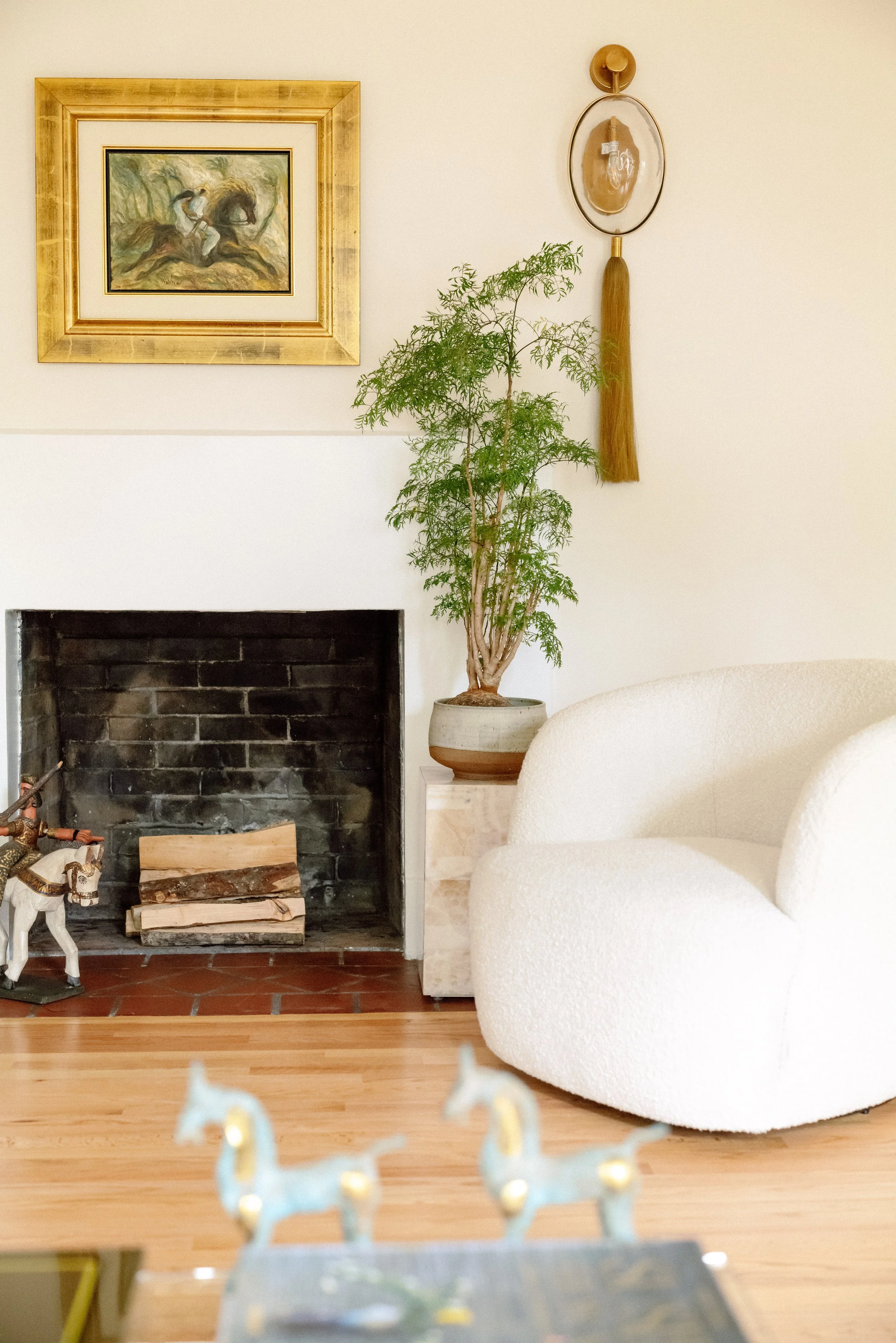 A cozy living room corner with a white rounded armchair, a potted plant next to a fireplace, a framed painting above the fireplace, and decorative items on the wall.