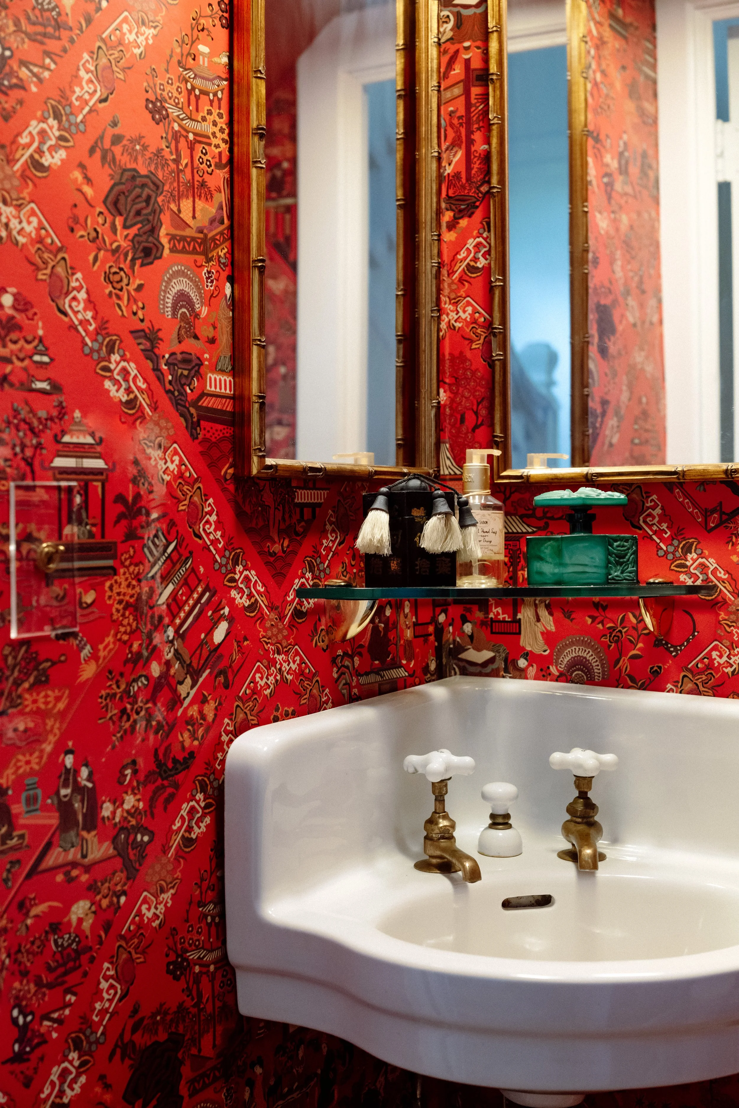 Close-up of a vintage white porcelain sink with brass faucets, set against red ornate wallpaper with Asian-inspired patterns and a mirror above, in a decorated bathroom.